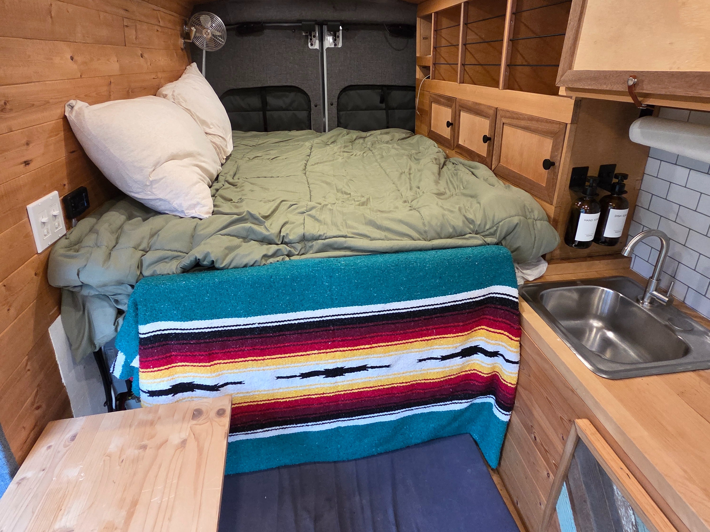 Inviting rv rental camper van interior with a comfortable bed, vibrant blanket, wood paneling, and a functional mini-kitchen.. Ford Transit 2015