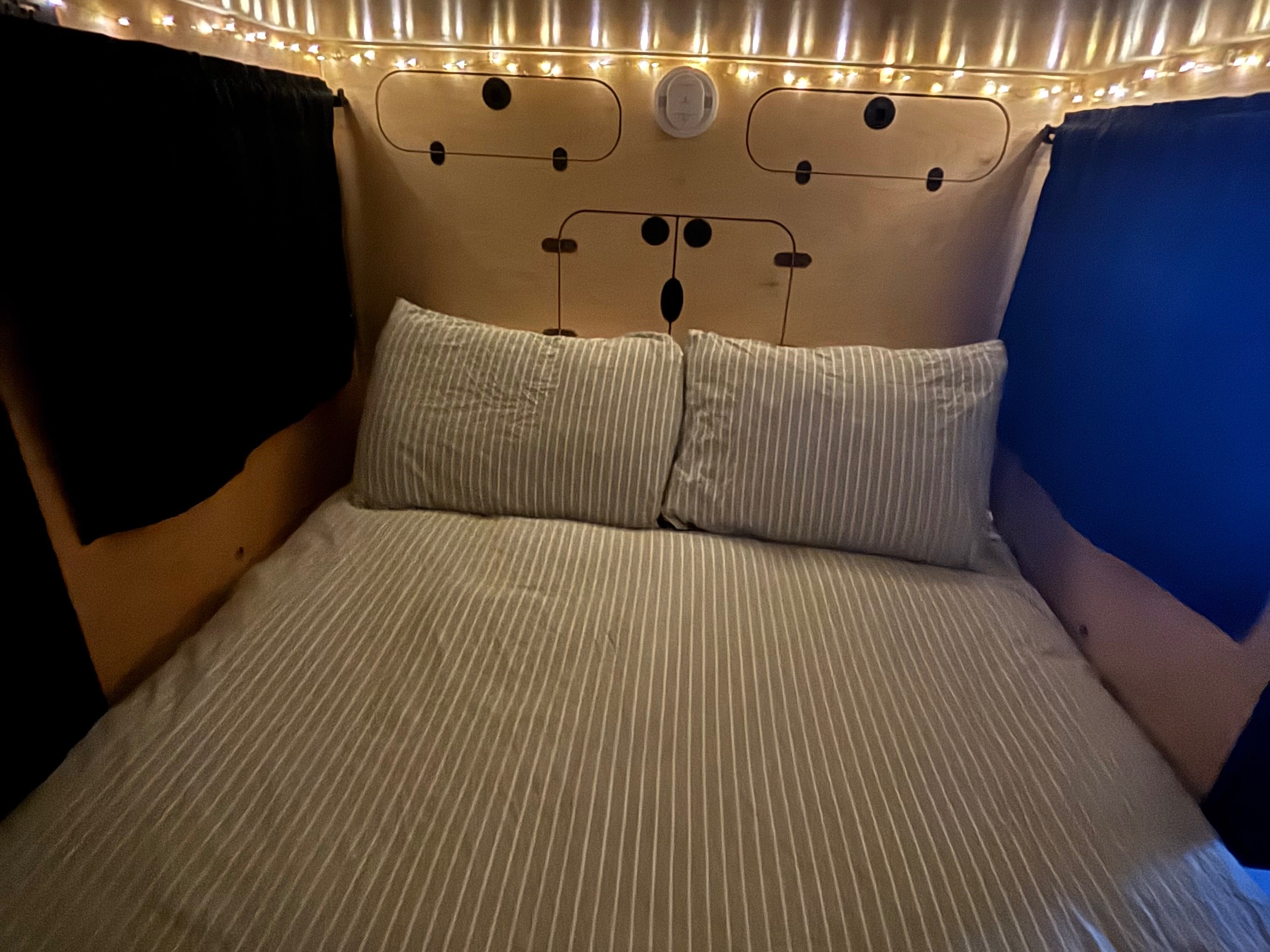 Cozy RV rental bed with striped bedding, string lights, and a unique bear-faced wooden panel overhead.. Hiker 5x8 Highway Deluxe 2025