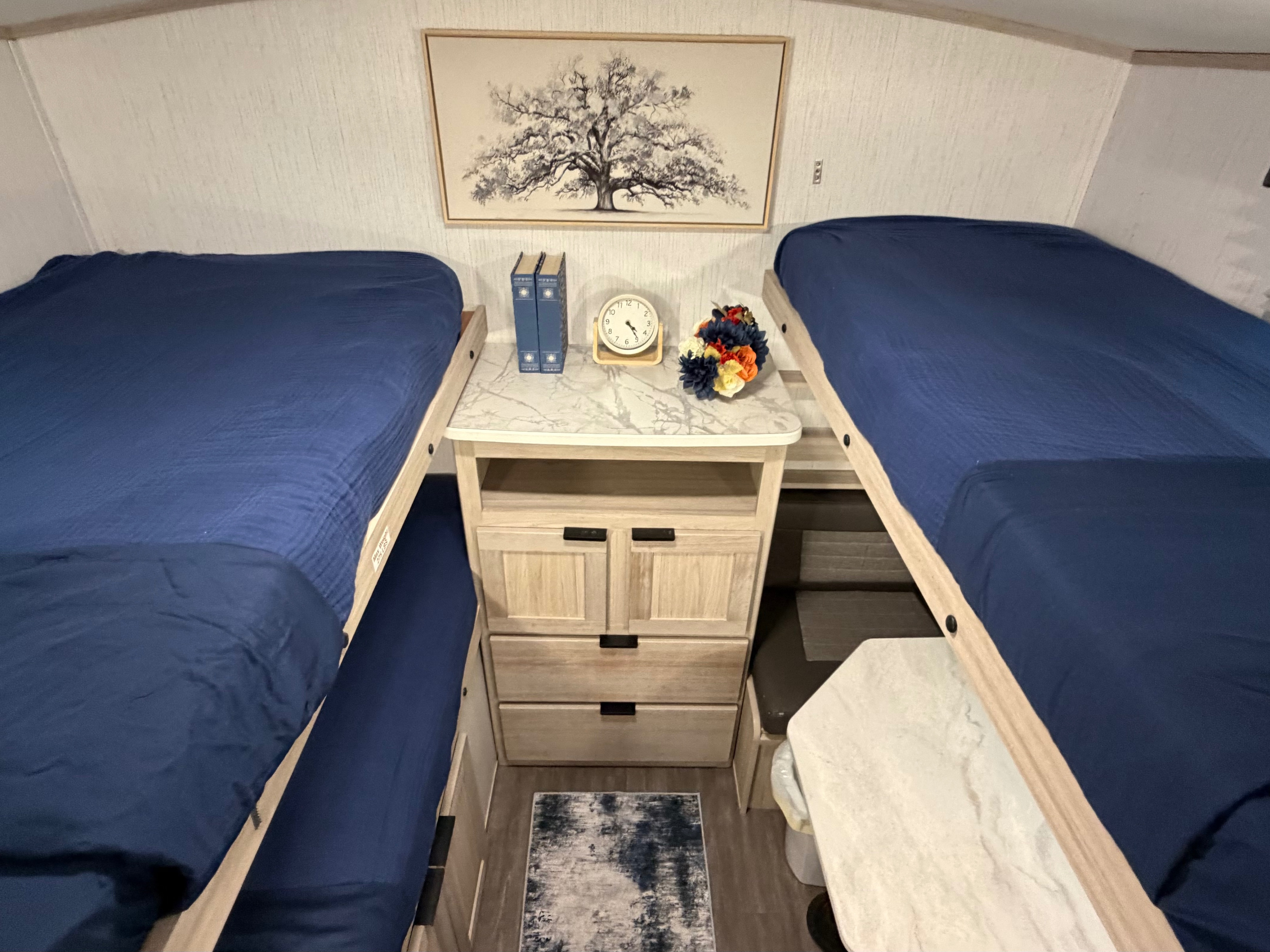 Cozy RV rental interior featuring twin bunk beds with blue bedding, a central nightstand, tree art, clock, and flowers.. Cruiser RV Shadow Cruiser 2022