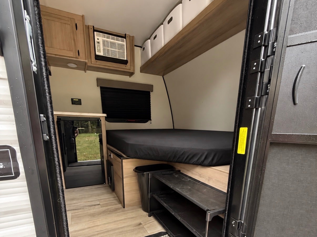RV rental interior showing a compact bedroom area with a bed, AC unit, mini-fridge, and overhead storage.. Keystone Coleman 13b 2027