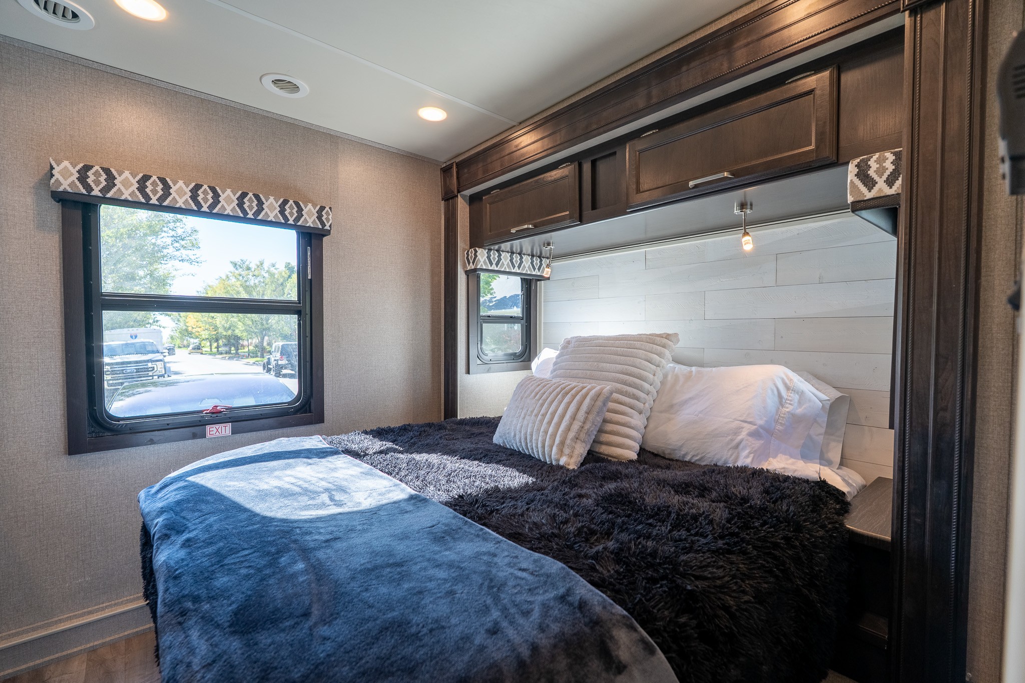 Cozy bedroom interior in an RV rental, featuring a bed with dark fuzzy bedding, pillows, windows with outdoor views, and overhead storage.. Jayco Greyhawk Prestige 2020