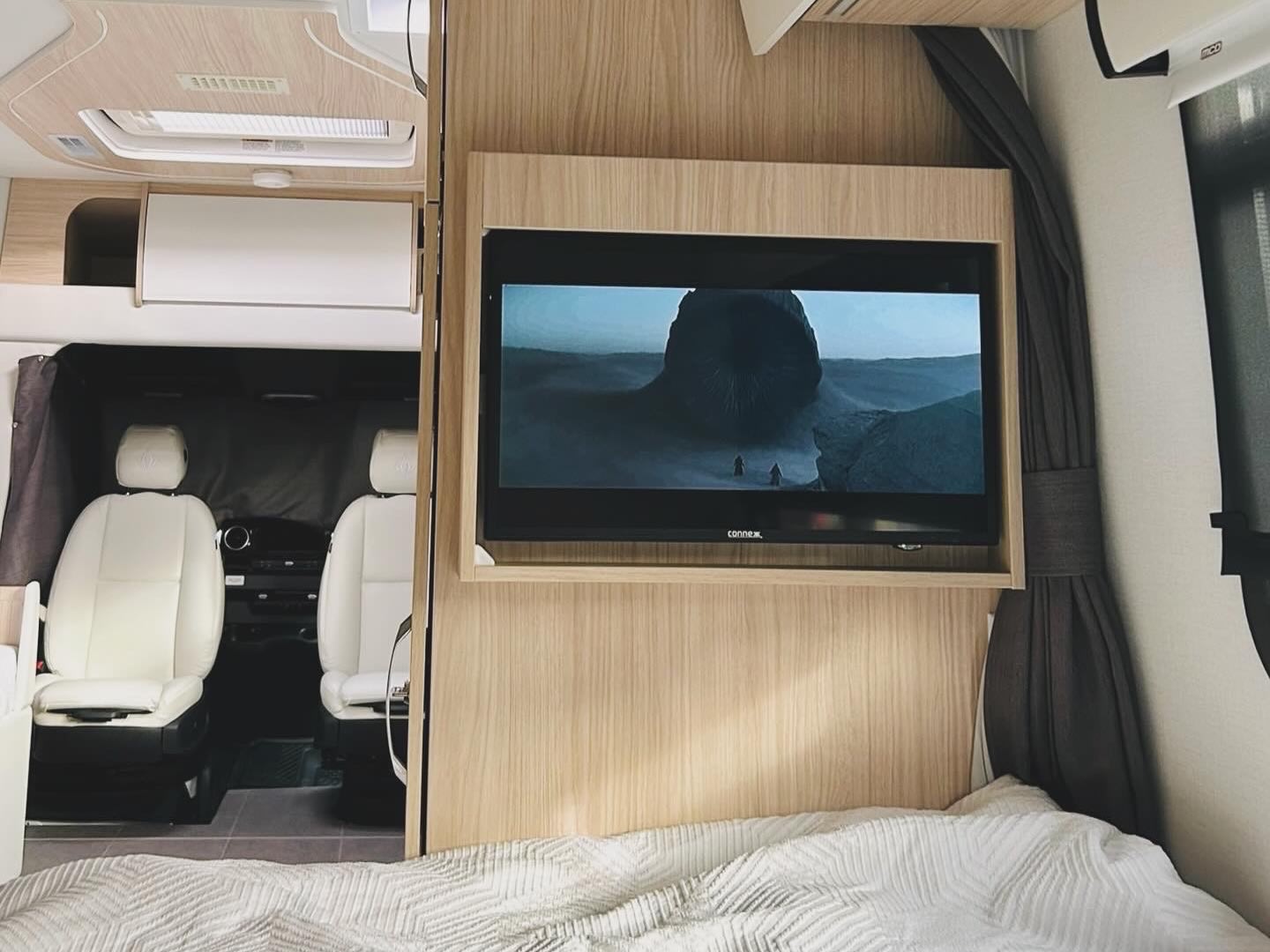 RV rental interior with a TV playing a movie, comfortable bed, and front seats, offering mobile comfort for your adventures.. Leisure Travel Vans Unity 2024