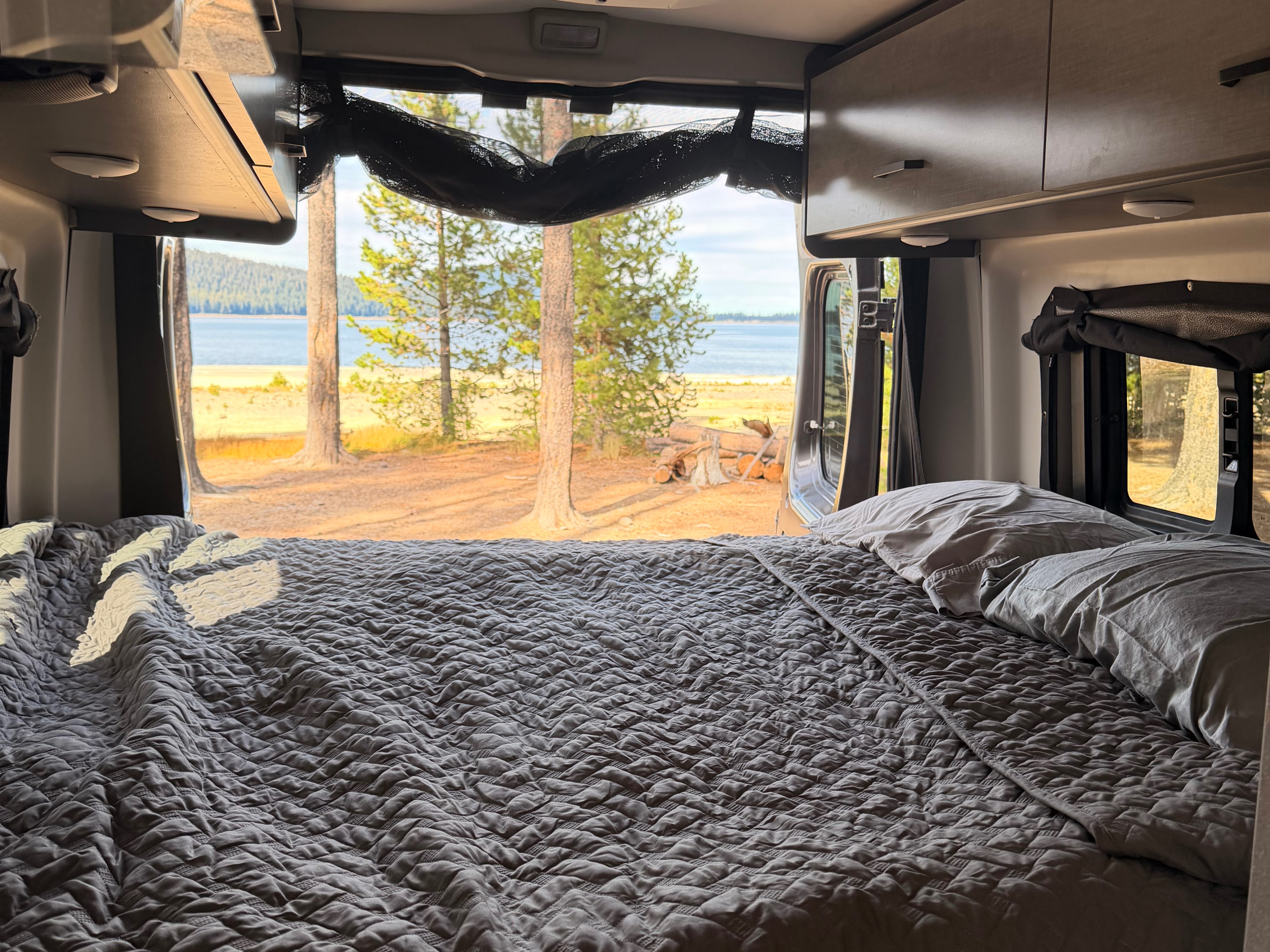 RV rental: Comfy bed with open back doors revealing a serene lake, forest, and sandy shoreline.. Thor Motor Coach Dazzle 2AB 2023