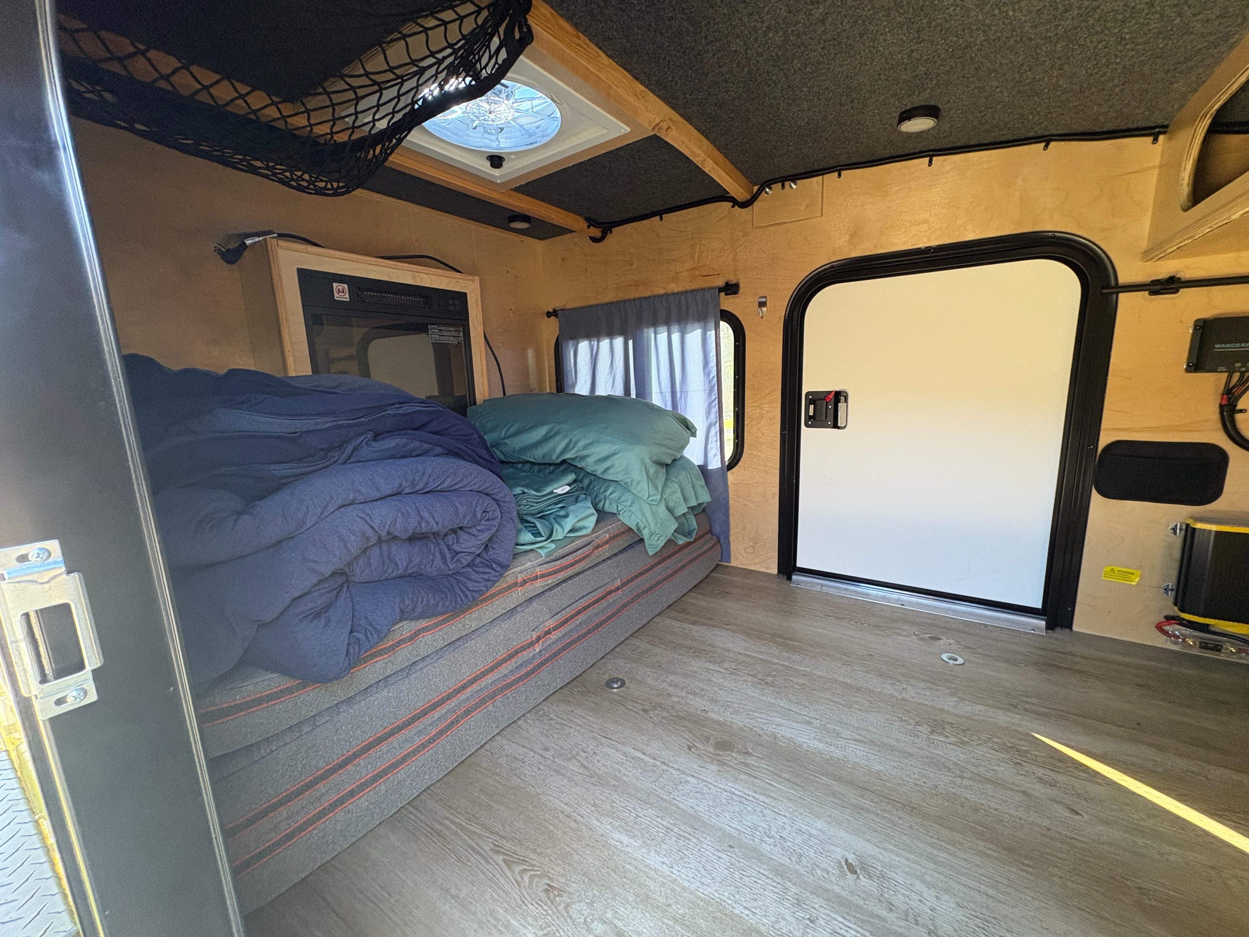 Inside an RV rental: a compact sleeping area with bedding, window, fan, and door, featuring wood walls & cozy design.. Outbound Trailers Outbound Extreme 2023