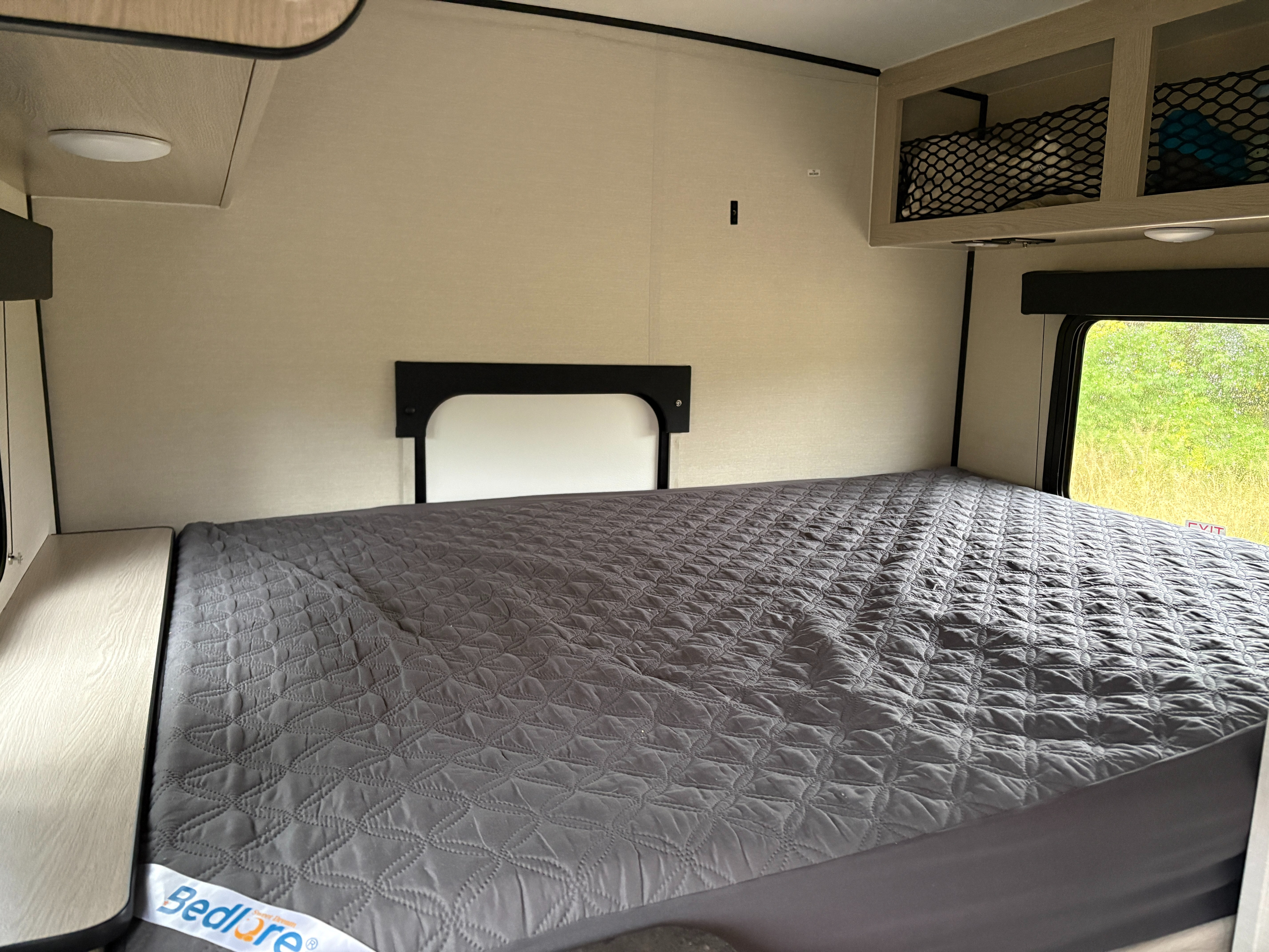 Experience comfort in this RV rental bedroom featuring a gray quilted bed, overhead storage, and a window view of lush nature.. Coachmen Apex Remote 16R 2023