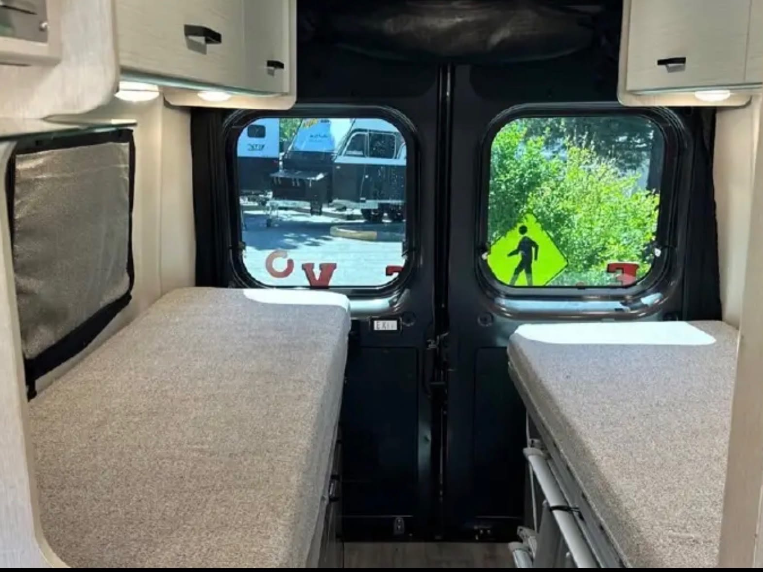 Cozy twin beds inside an RV rental, offering a view out the back windows to trees and a pedestrian crossing sign.. Thor Dazzle 2024