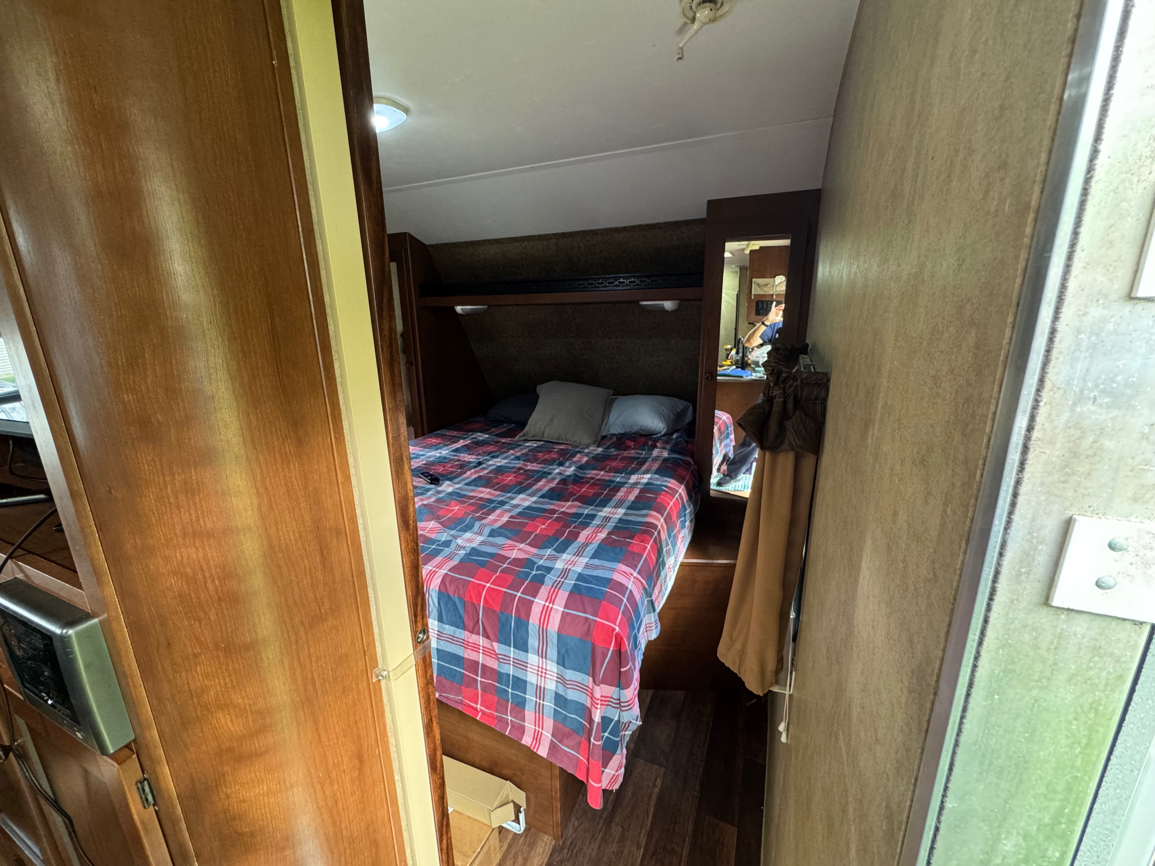 Inside an RV rental: a compact bedroom with a red and blue plaid comforter, wooden walls, and a mirror showing a person.. Aspen Trail Aspen Trail Trailer 2012