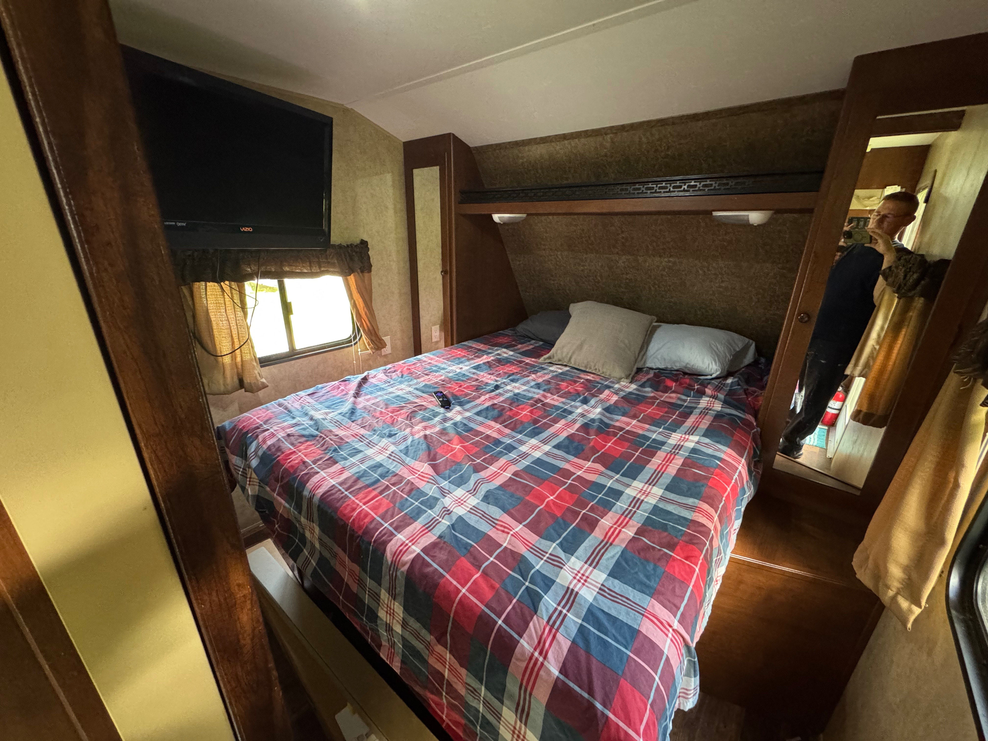 RV rental: a cozy bedroom with plaid bed, TV, window, and overhead storage, perfect for road trip comfort.. Aspen Trail Aspen Trail Trailer 2012