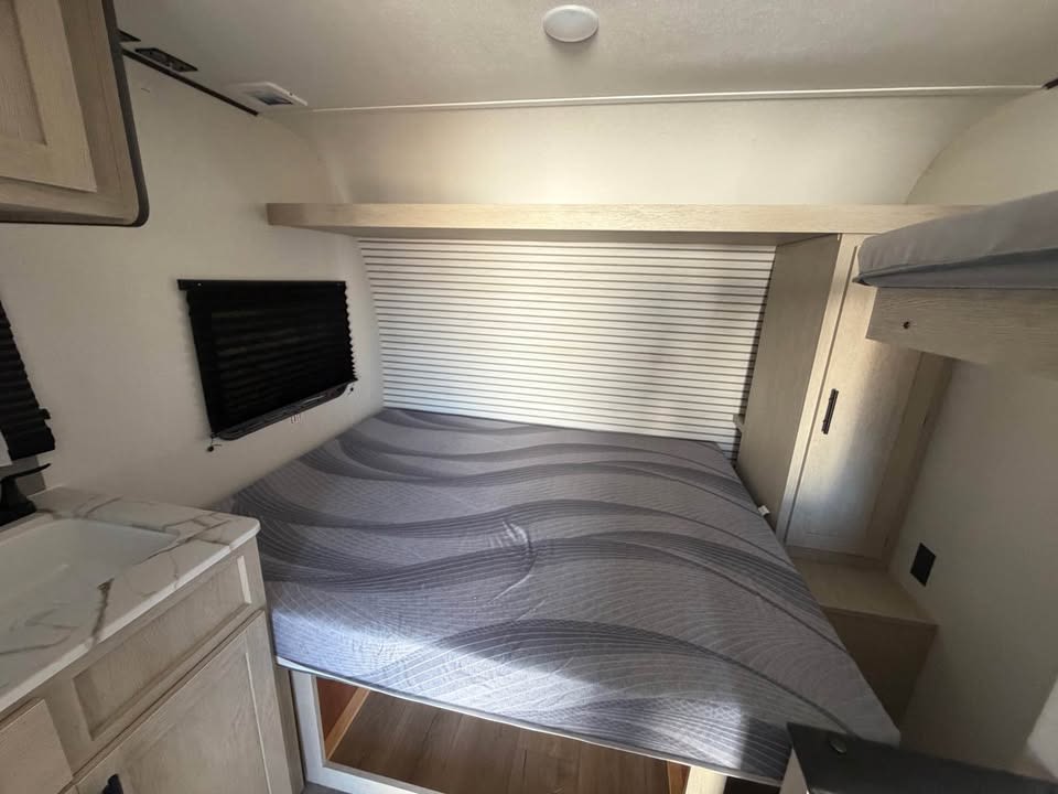 RV rental bedroom: main bed with gray mattress, upper bunk, small sink vanity, and window with dark blind.. Coachmen Shasta 2027