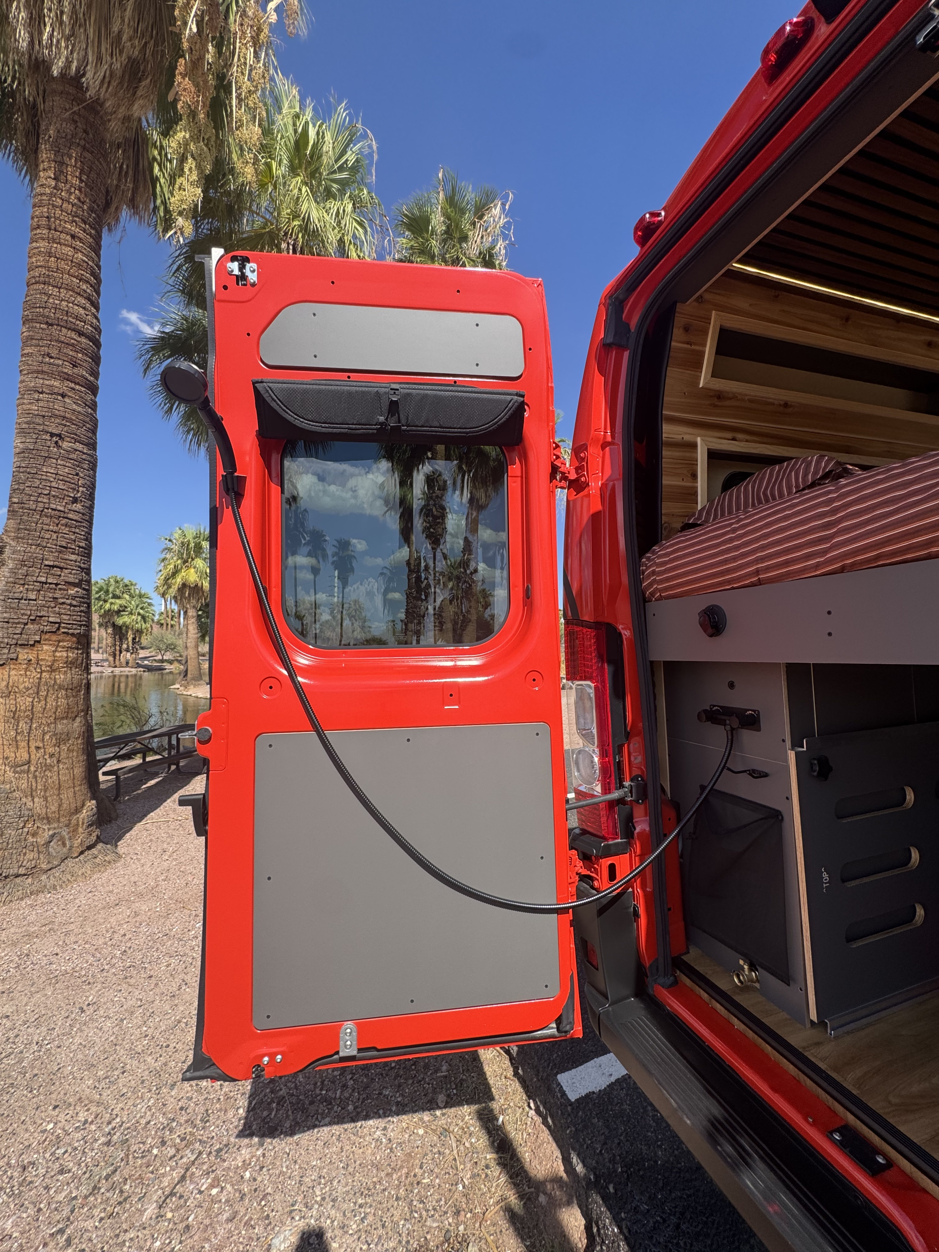 Red rv rental van with open back doors, revealing a comfy bed and outdoor shower, overlooking a serene palm-lined lake.. Ram Promaster 2023