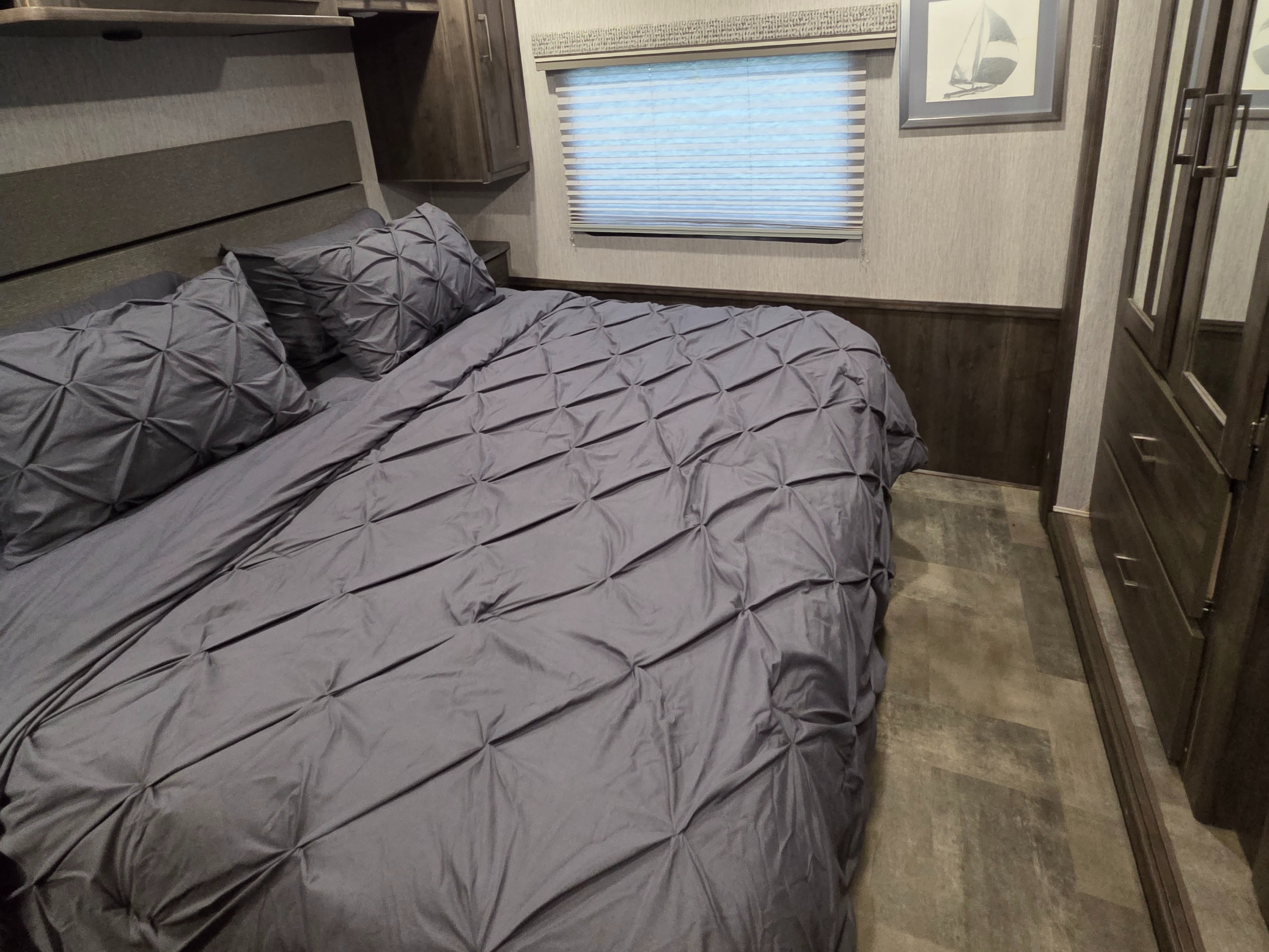 Cozy RV rental bedroom featuring a grey pintuck bed, window with blinds, wood cabinetry, and sailboat art on the wall.. Forest River Coachmen Pursuit 2022