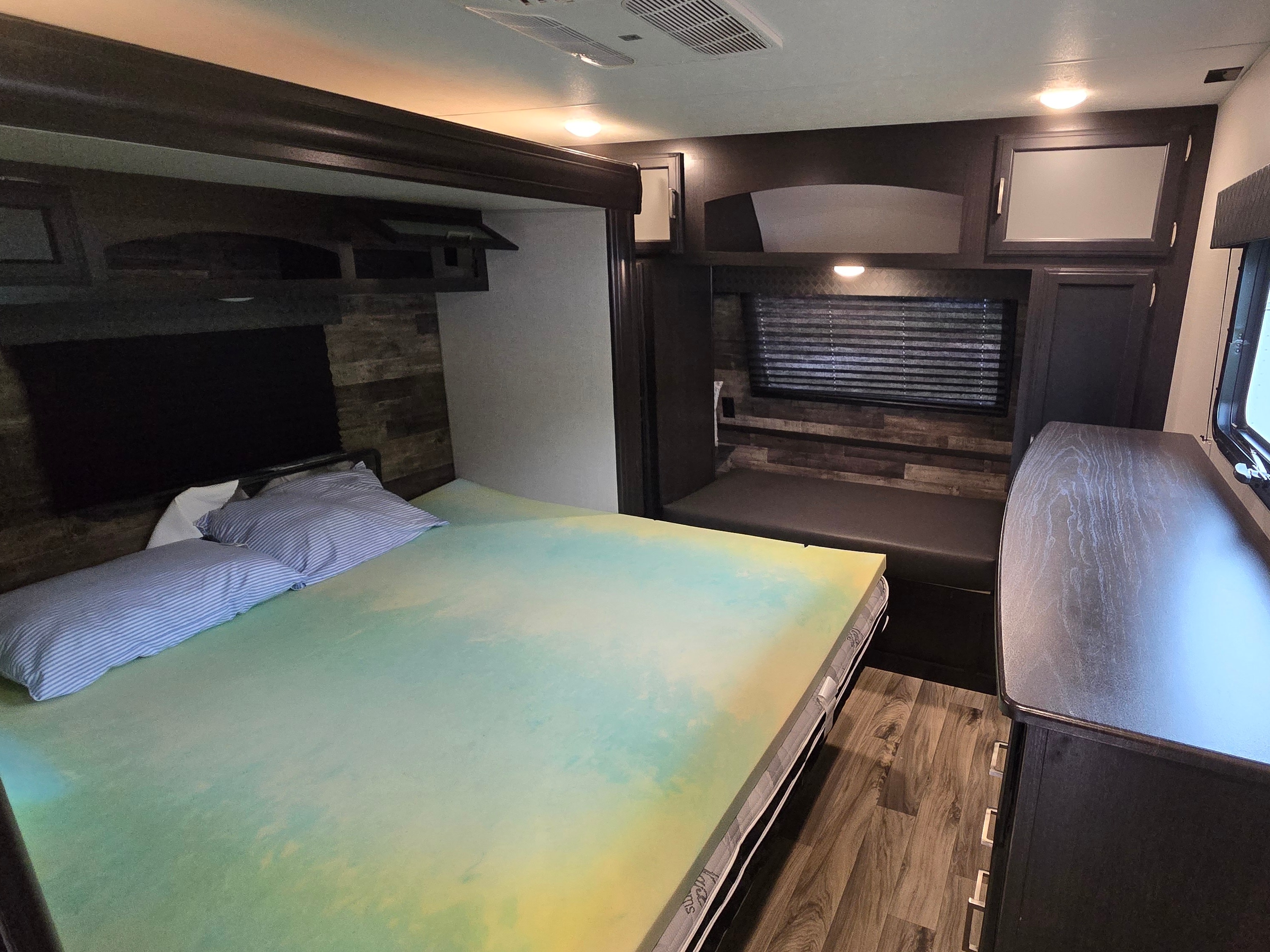 RV rental bedroom shows a compact interior with a bed, colorful topper, dark wood cabinetry, window with blinds, and bench seat.. Sport Trek Touring 2021