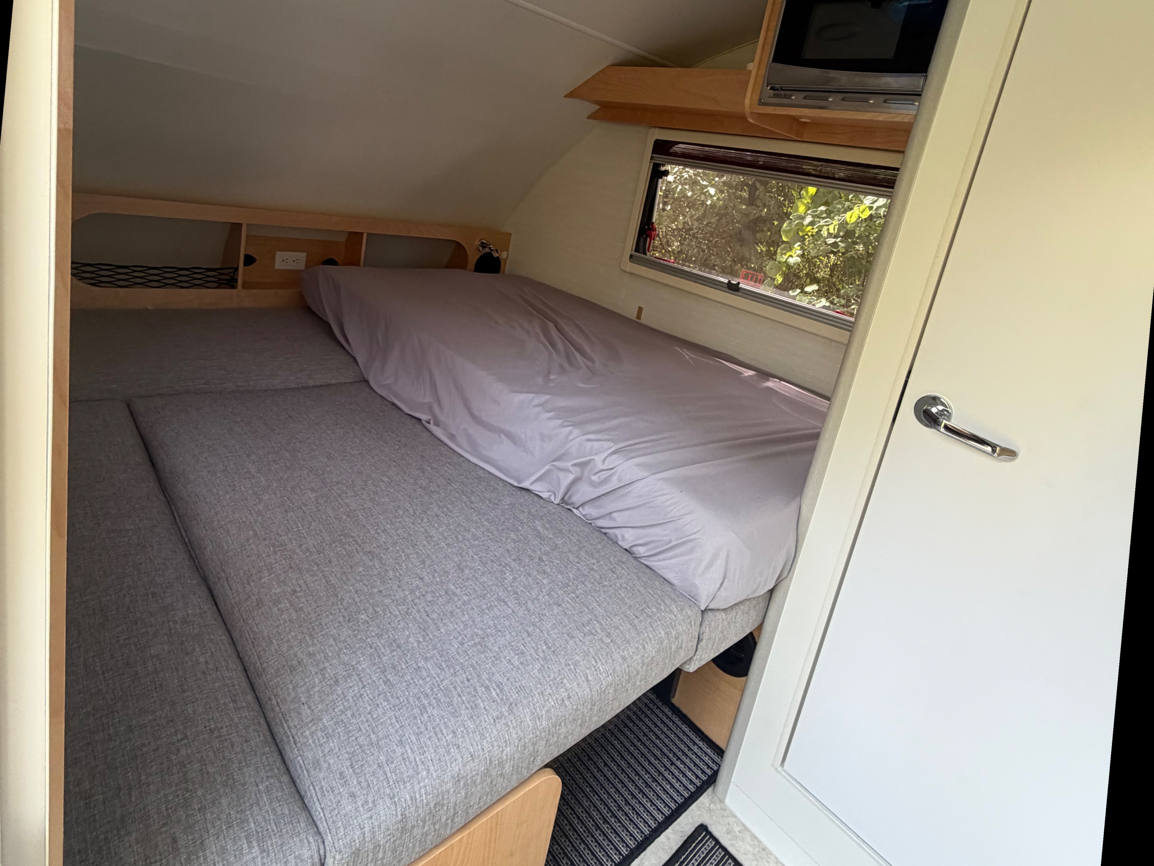 The compact interior of an rv rental features a bed, a window with foliage view, microwave, and storage cubbies.. nuCamp T@B 2026