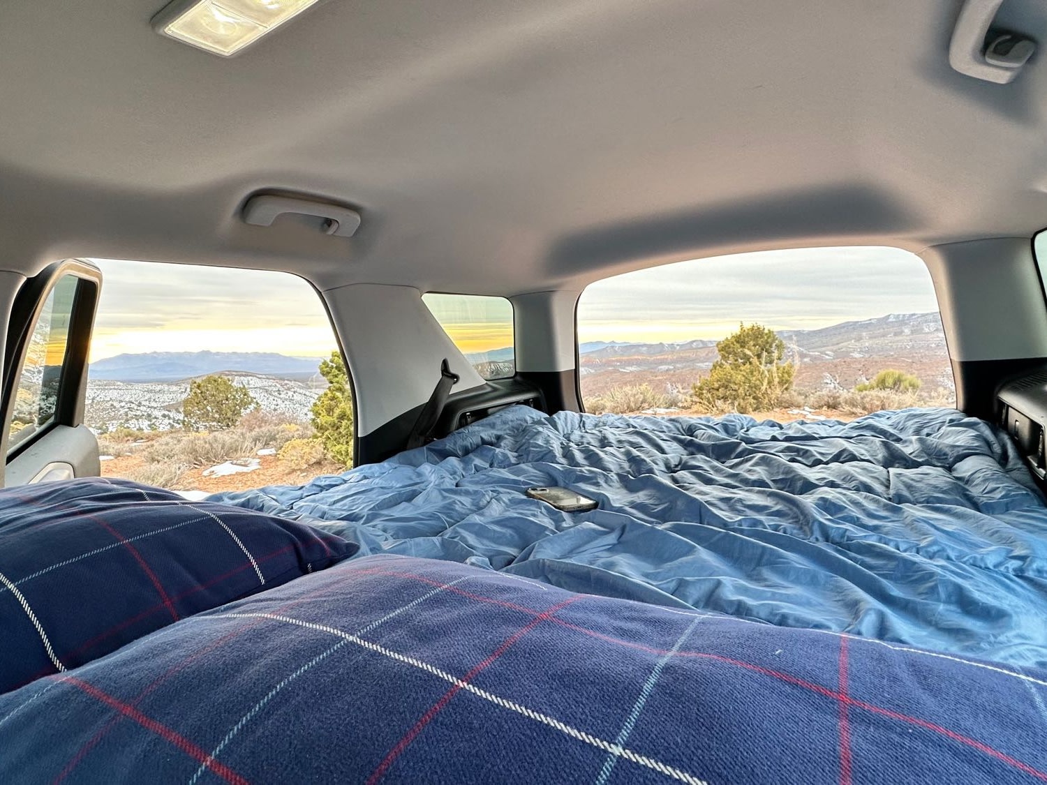 RV rental offers a cozy car camping bed with blue comforter & plaid pillows, facing scenic snowy mountains at sunset.. Ford Bronco Sport 2022