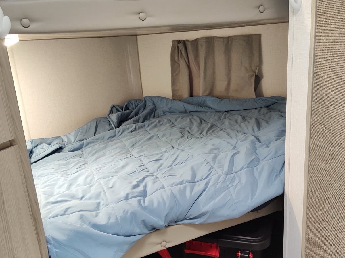 A compact sleeping area in an RV rental with a blue quilted comforter, headboard, and under-bed storage bins.. Forest River Wildwood FSX 2024