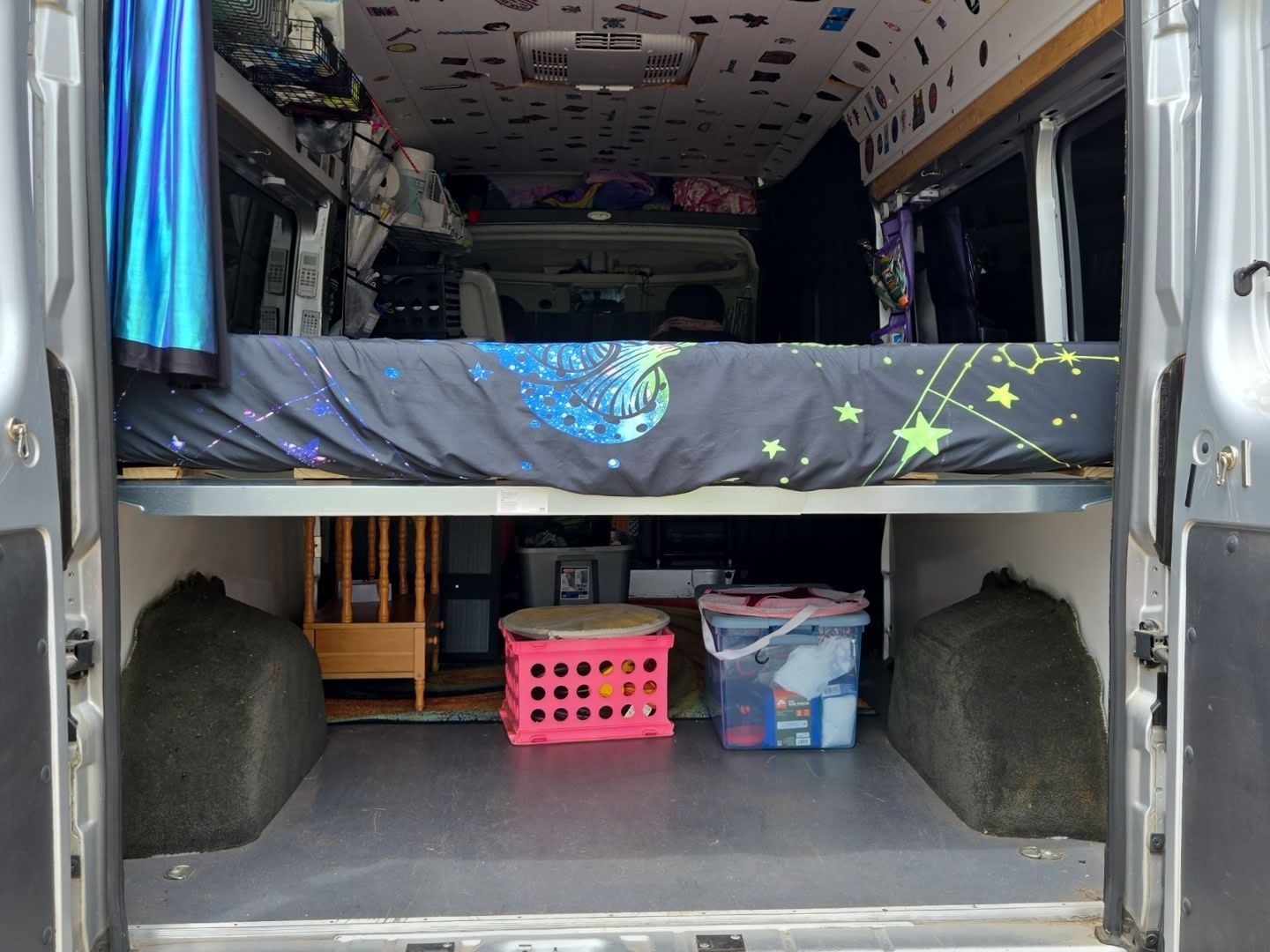 Rear interior of an RV rental with a galaxy-themed bed over under-bed storage, featuring a stool and bins for camper life.. Dodge Ram Promaster 2017