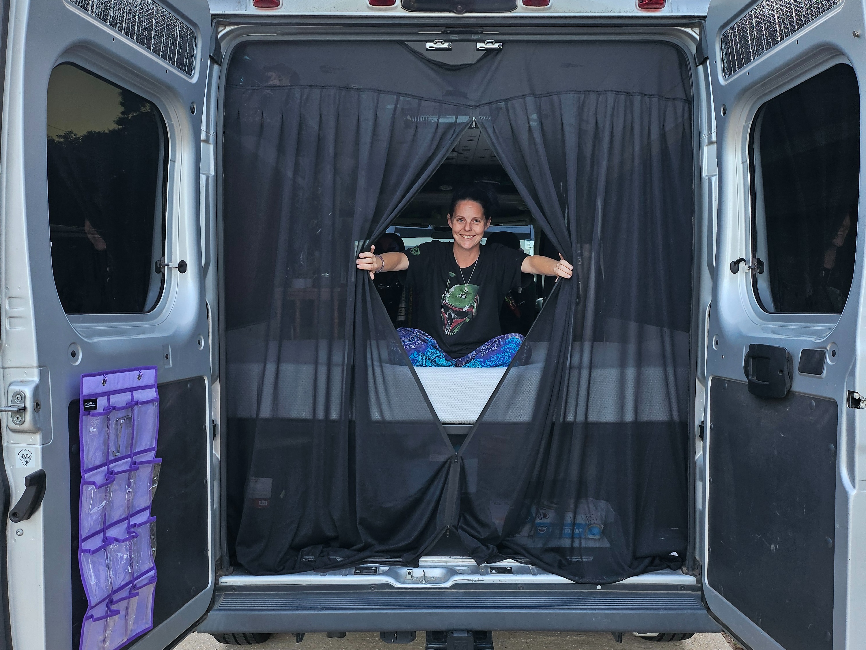 Smiling woman inside an RV rental, peeking through a mesh screen in the open back doors. Ideal for camper van adventures!. Dodge Ram Promaster 2017