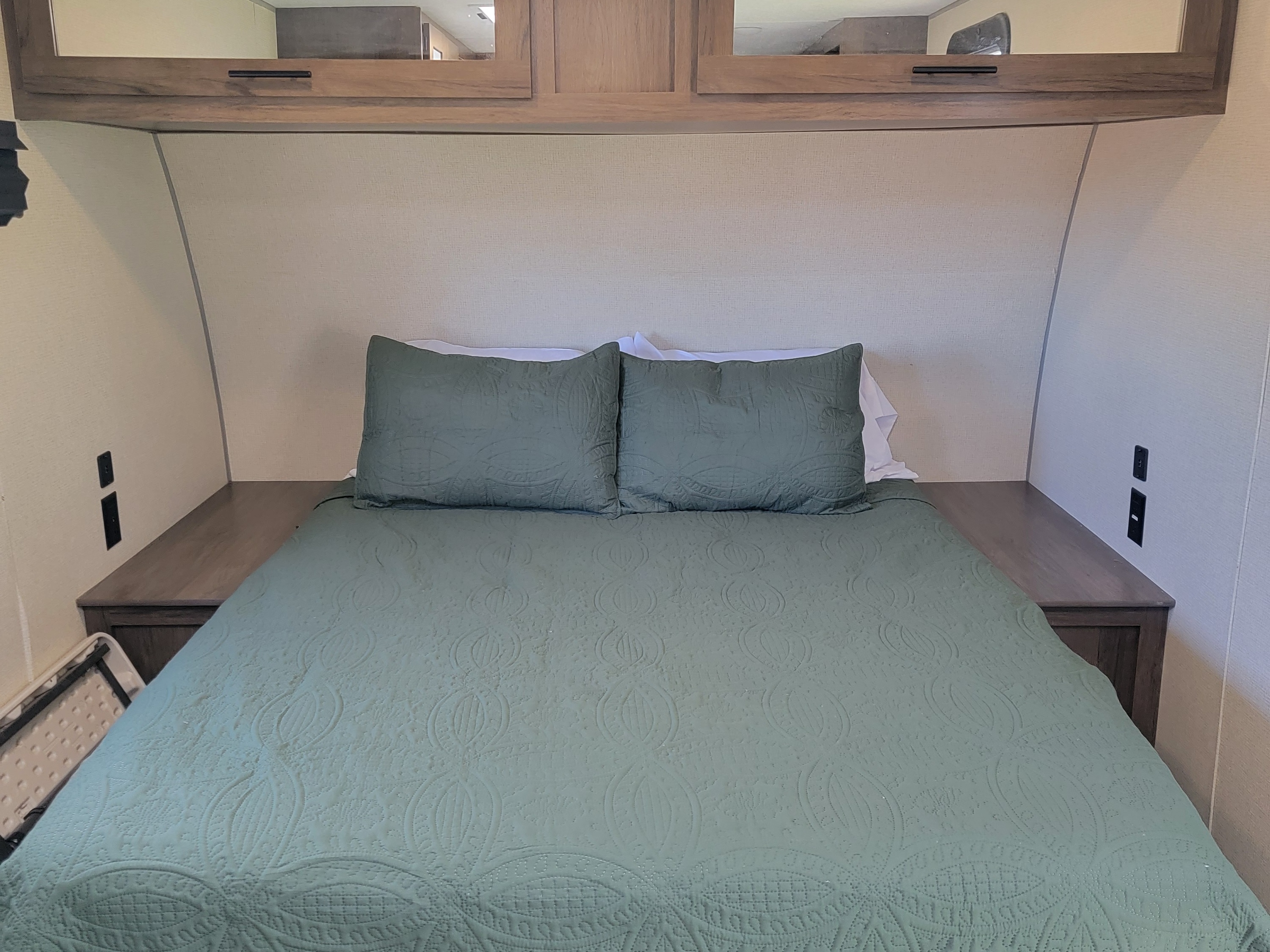 Cozy RV rental bedroom features a green quilted bed, built-in nightstands with outlets, and overhead cabinets.. Forest River Avenger 2020