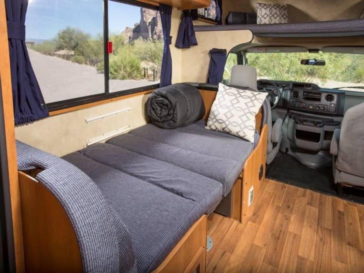 Cozy RV rental interior showing a bed with pillows, a window revealing a desert landscape, and the driver's cabin. Ideal for travel.. Thor Majestic 2023