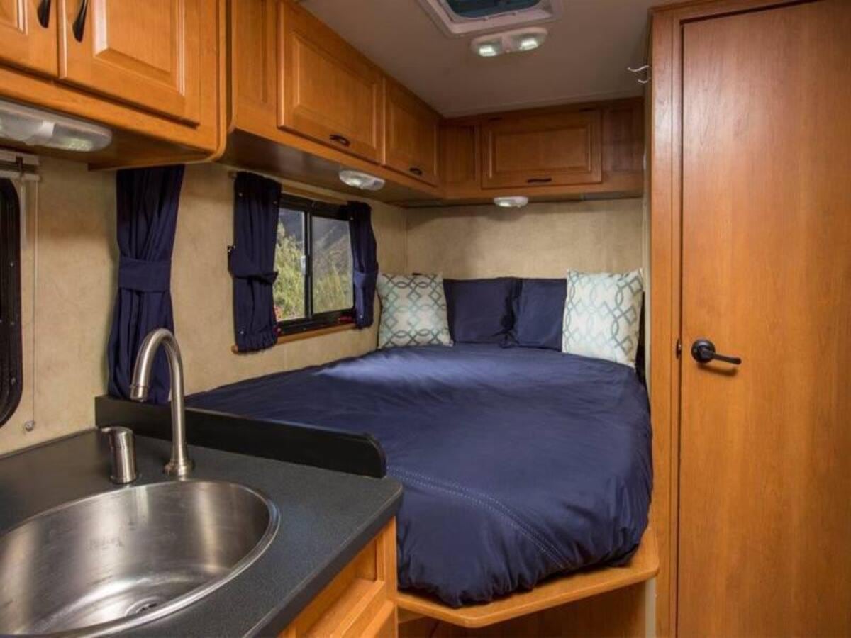 Inside an RV rental, a comfy bed with blue bedding next to a small kitchen sink and wood cabinets, offering cozy travel.. Thor Majestic 2023