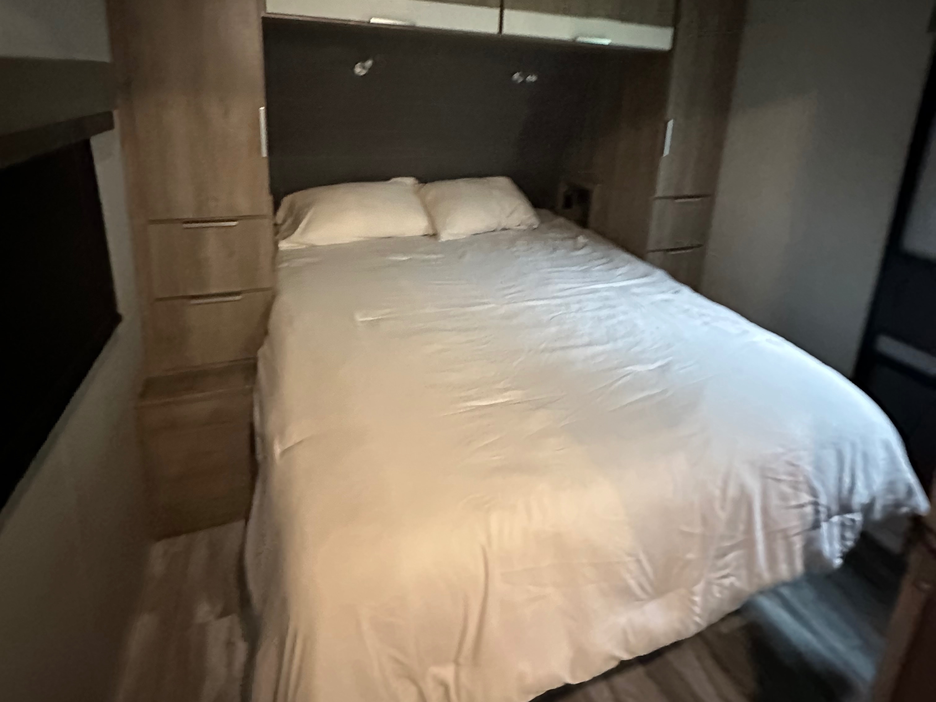Cozy RV rental bedroom features a queen bed with white linens, flanked by storage cabinets and overhead compartments.. Grand Design Imagine 2021