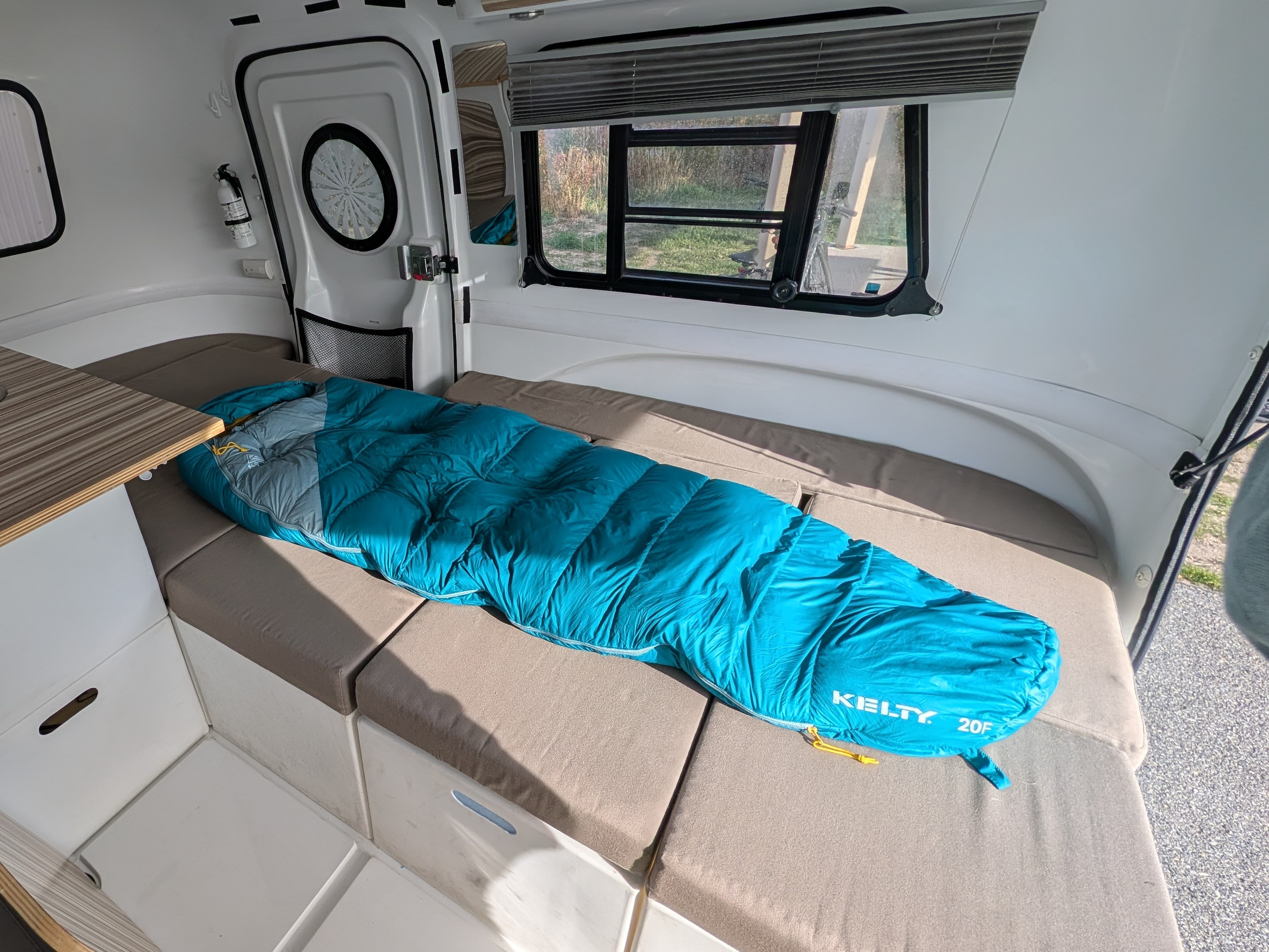 This rv rental's cozy campervan sleeping area features a Kelty sleeping bag on cushioned benches, with windows and a fire extinguisher visible.. Happier Camper HC1 2016