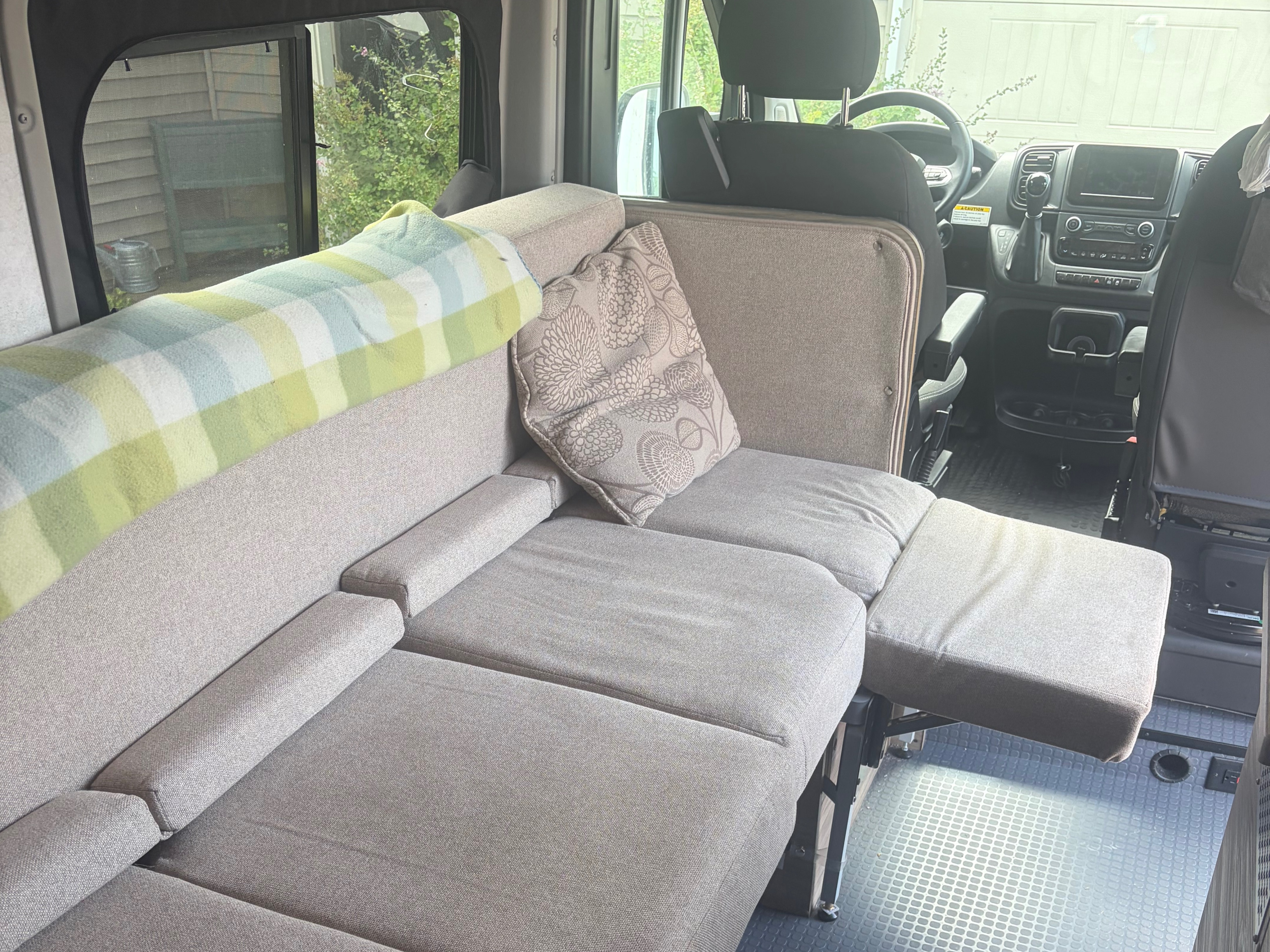 Spacious rv rental interior, showing a versatile light gray sofa/bed with a plaid blanket, pillow, and driver's area.. Winnebago Solis 2025