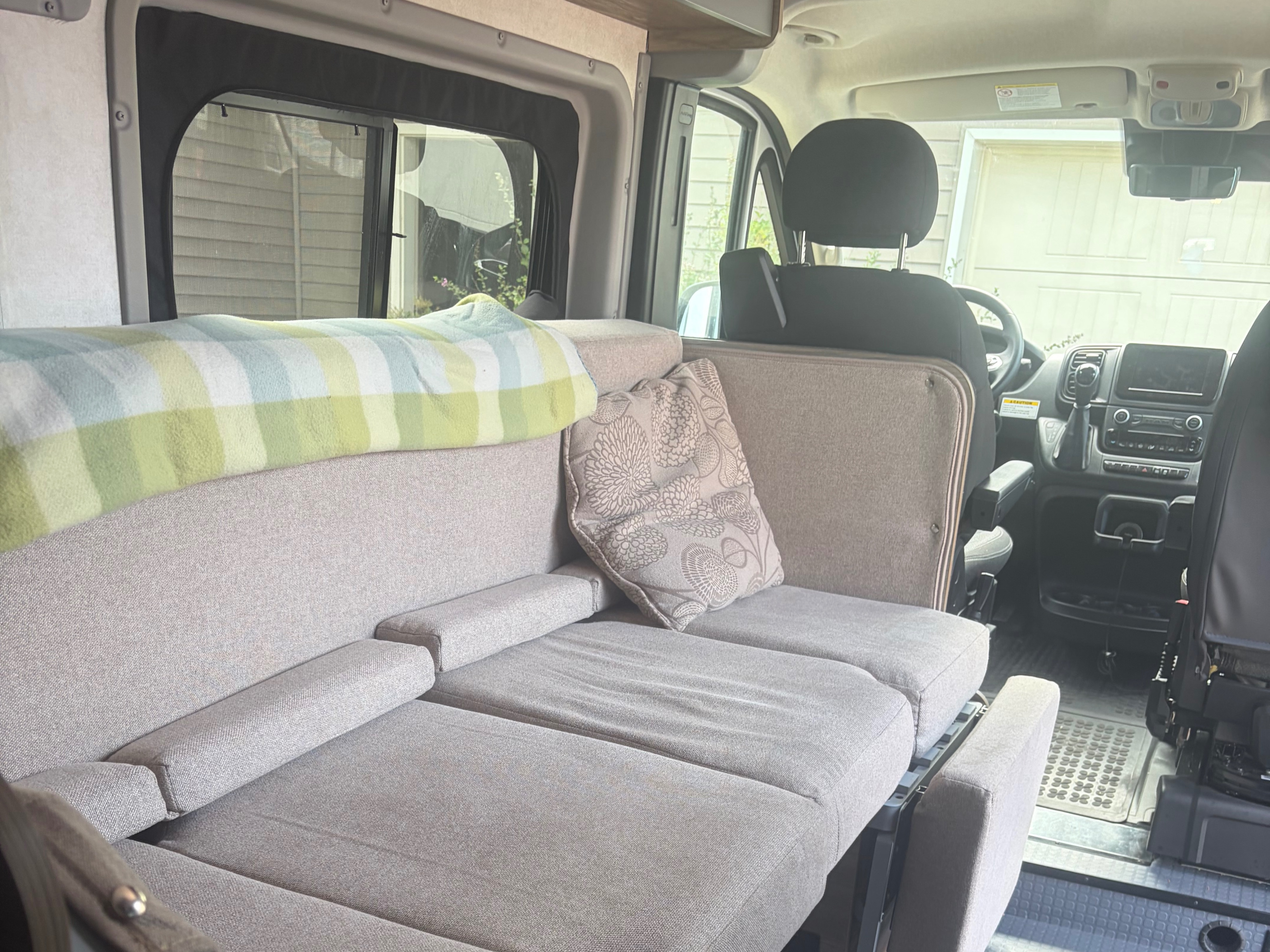 Inside this RV rental, a light-colored sofa converts to a bed, topped with a plaid blanket & floral pillow, facing the cockpit.. Winnebago Solis 2025