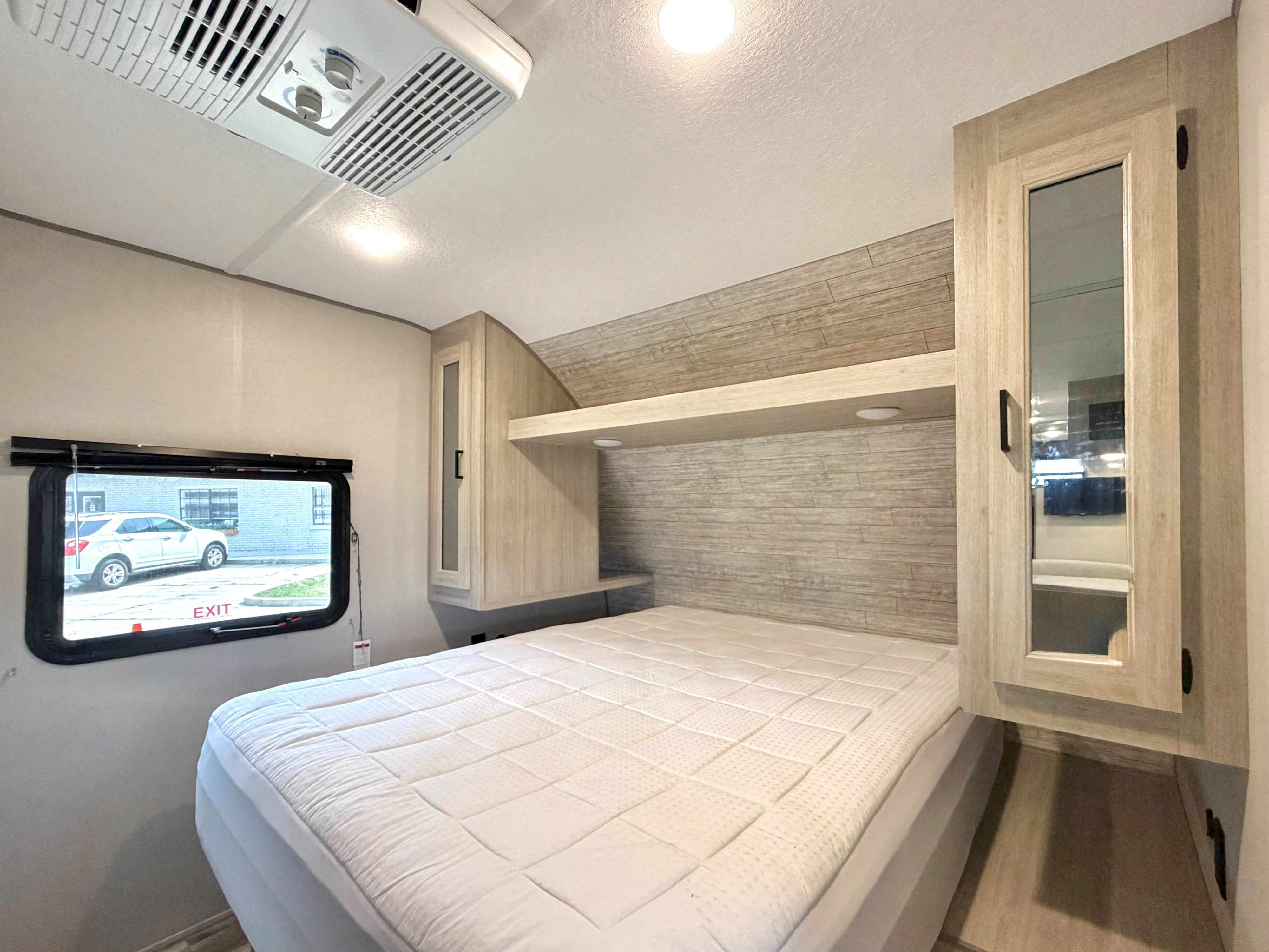 RV rental bedroom featuring a white quilted bed, overhead AC unit, emergency exit window, and modern light wood cabinetry with storage shelf.. Coachmen Catalina Summit 261BH 2025