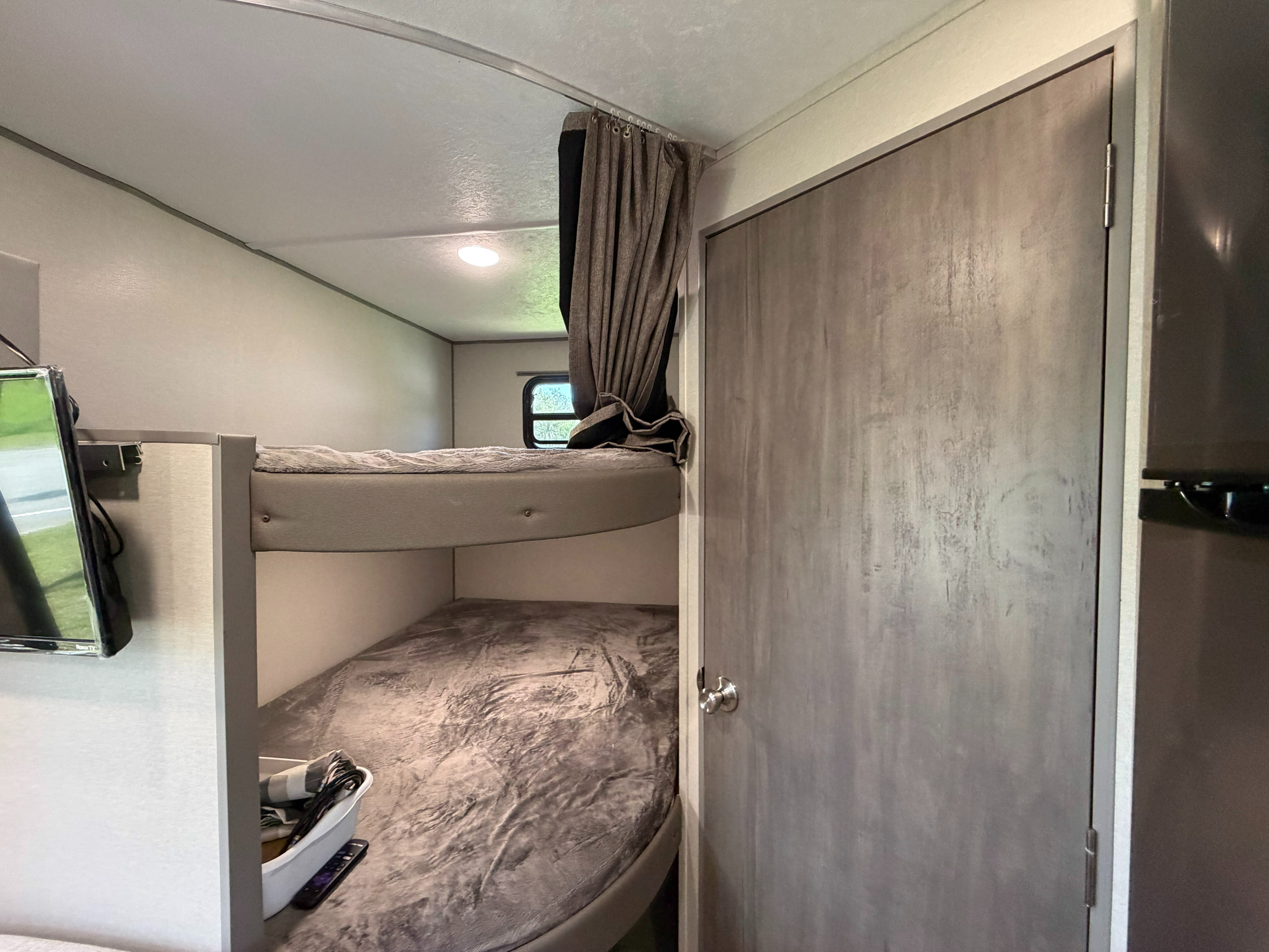 This RV rental features bunk beds with gray bedding, a small window with curtains, and a door, all in neutral tones.. Coachmen Catalina Summit 261BH 2025