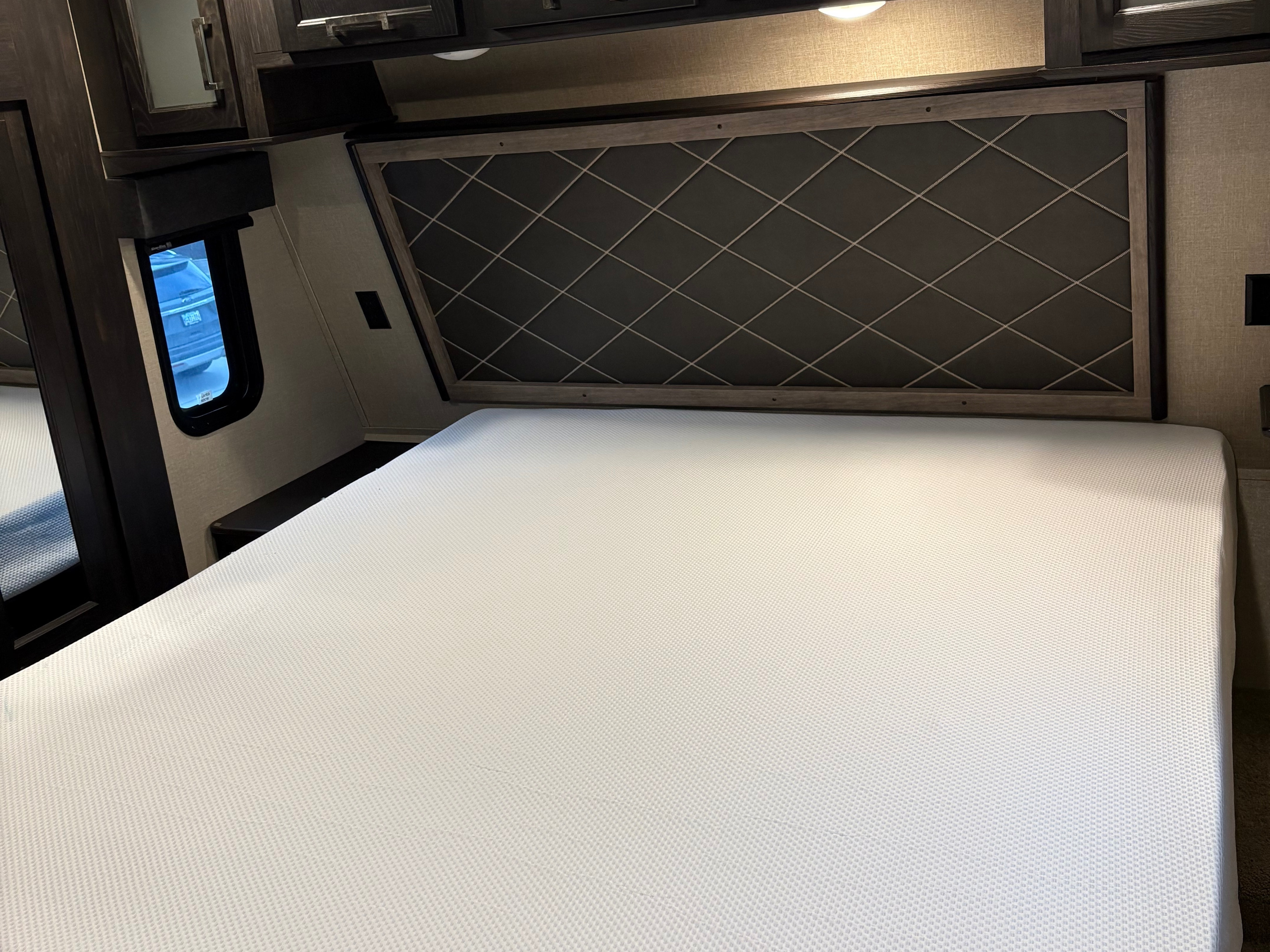 RV rental bedroom: a clean white mattress, quilted headboard, window view, and overhead cabinets.. Keystone Fuzion 2019