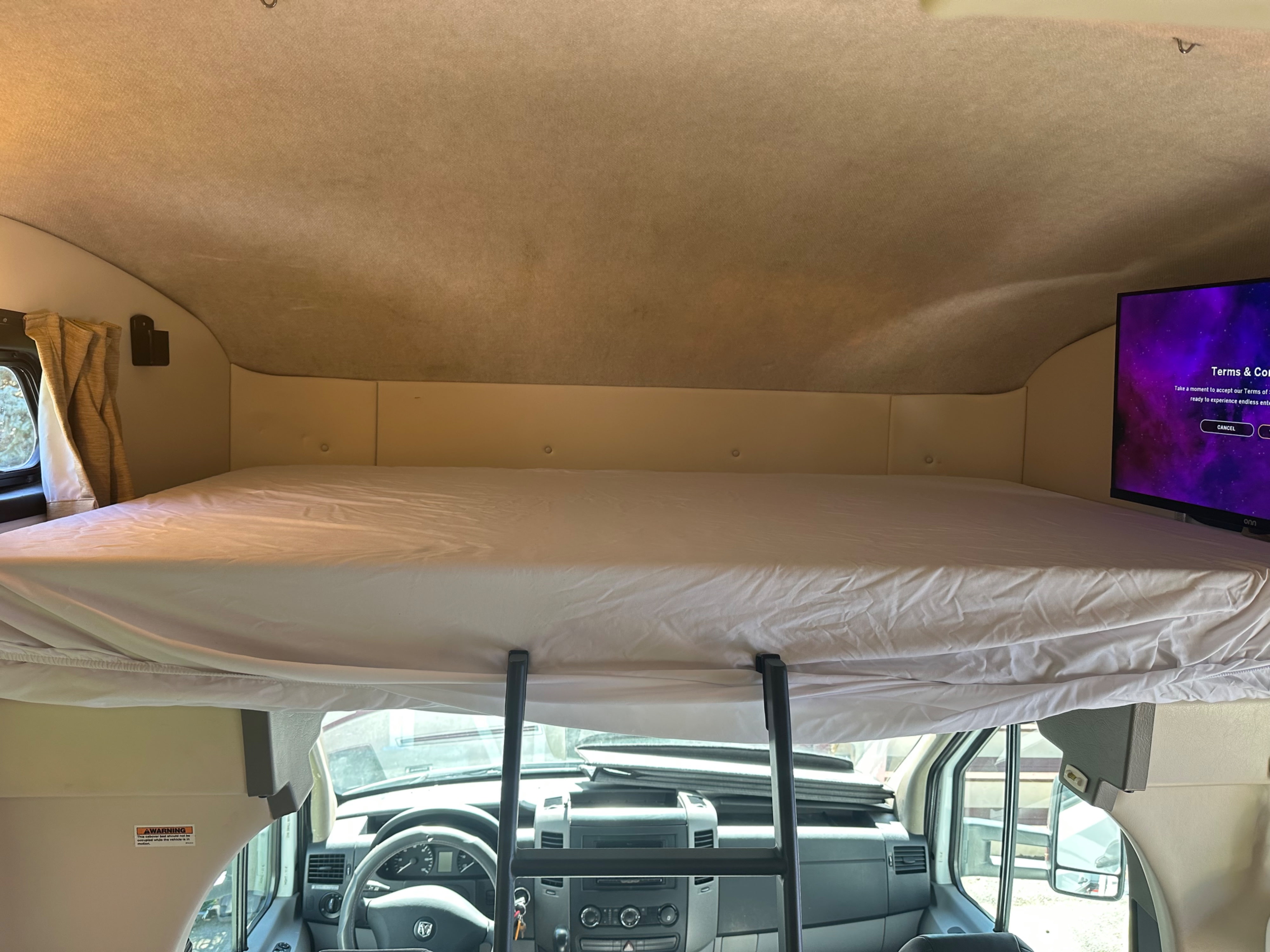Overhead bunk bed with white sheets and access ladder in an RV rental, showing the driver's cabin below and a TV.. Fleetwood Other 2010
