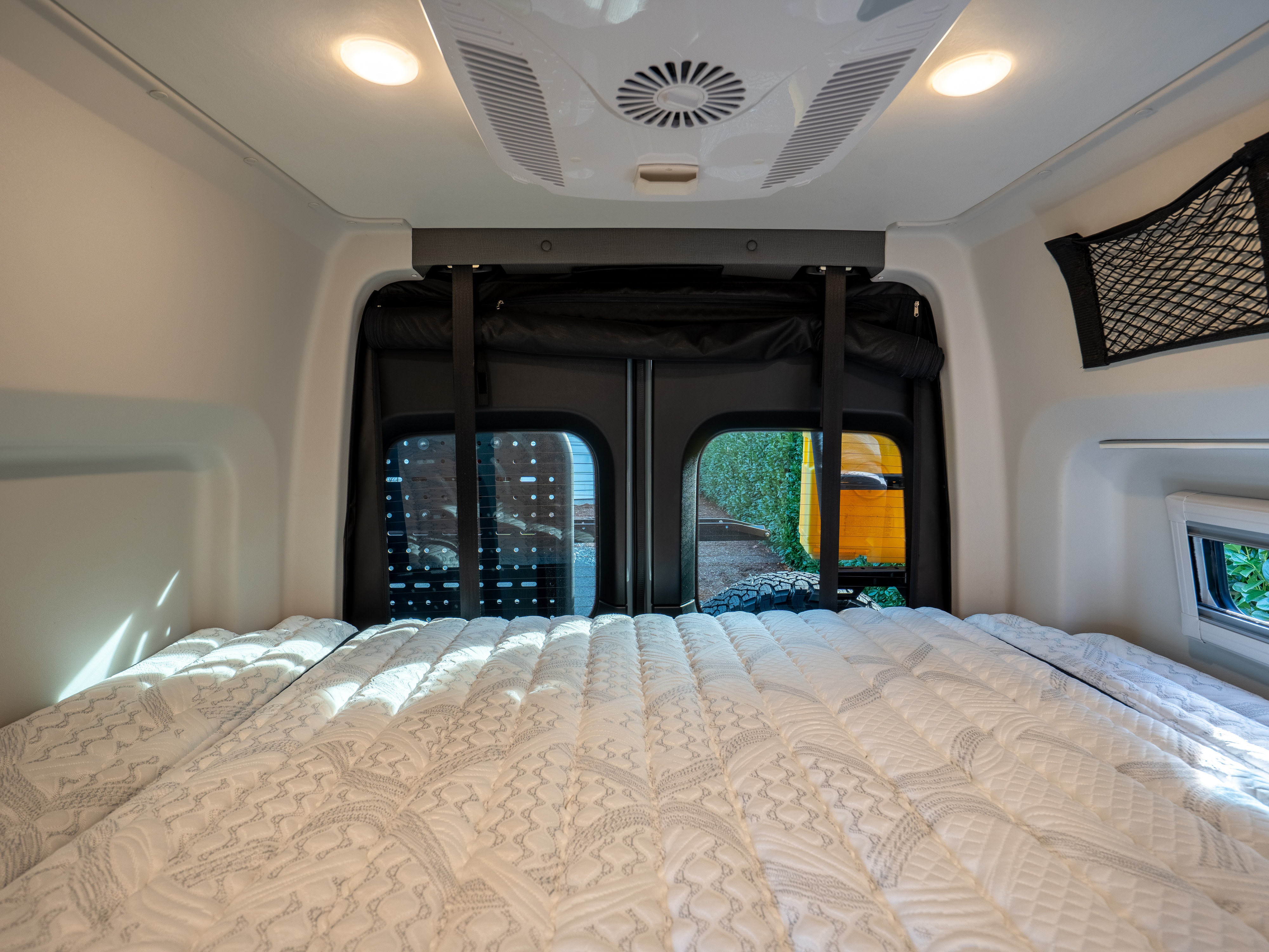 Inside a spacious RV rental: a comfortable bed, rear doors with outdoor views, overhead lighting, and AC unit.. Tiffin Motorhomes GH1 2025