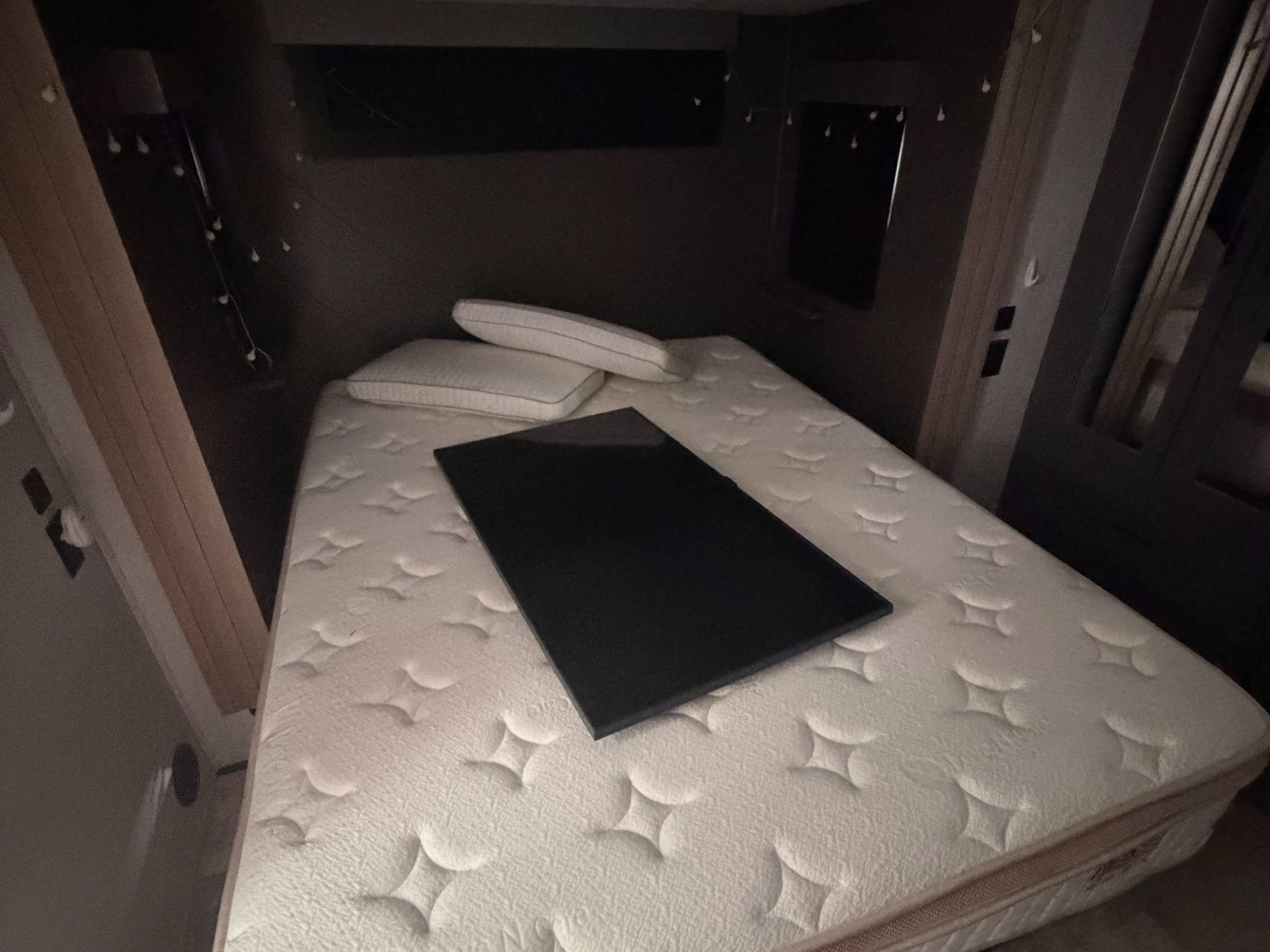 Compact RV rental bedroom with a mattress, pillows, and a black flat object. Cozy sleeping area with string lights.. Grand Design Momentum M-Class Toy Hauler 2023