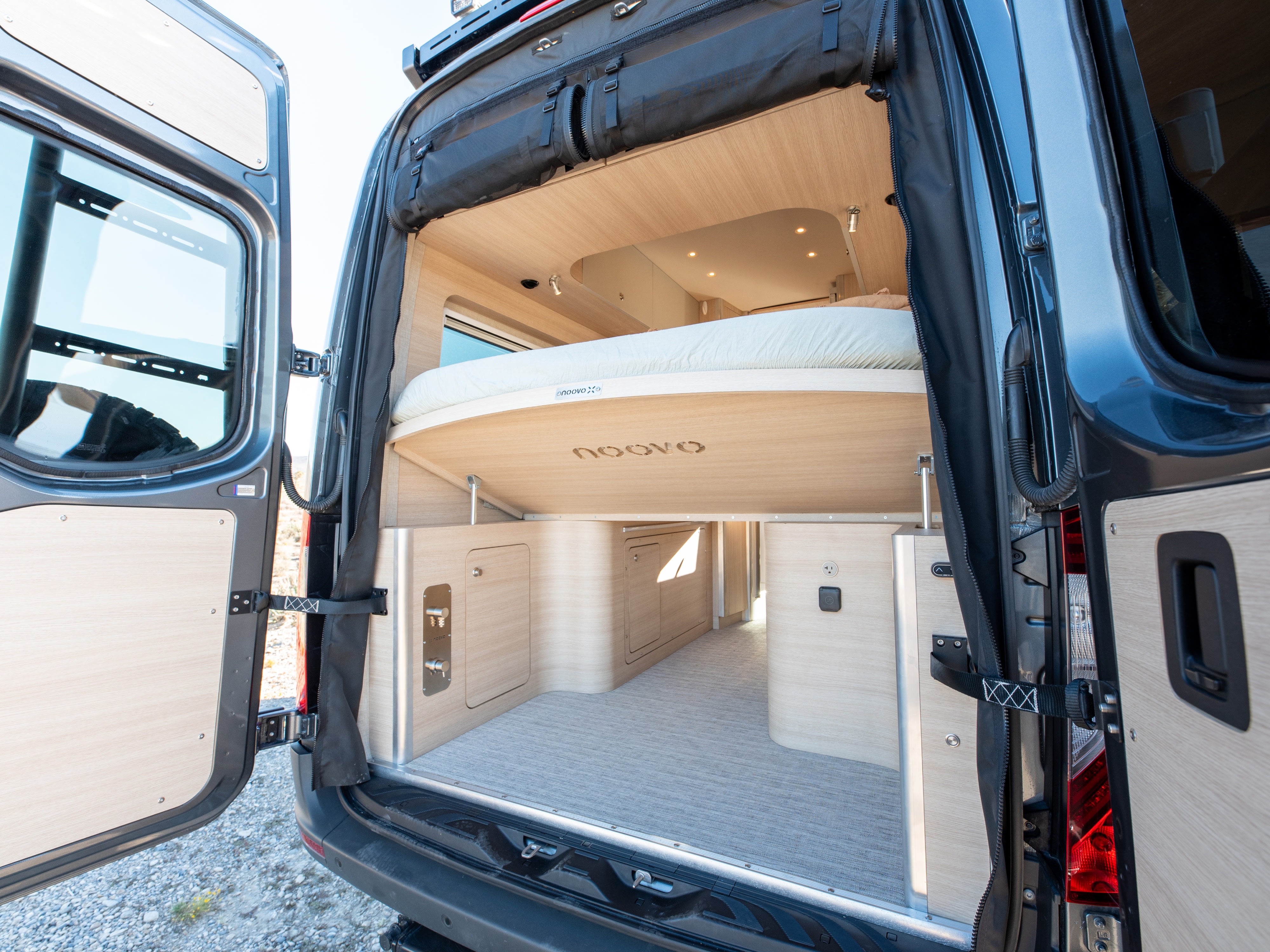 RV rental: Rear view into a modern camper van's interior, showing a raised bed above storage, with light wood finishes.. Noovo Noovo Next 2026