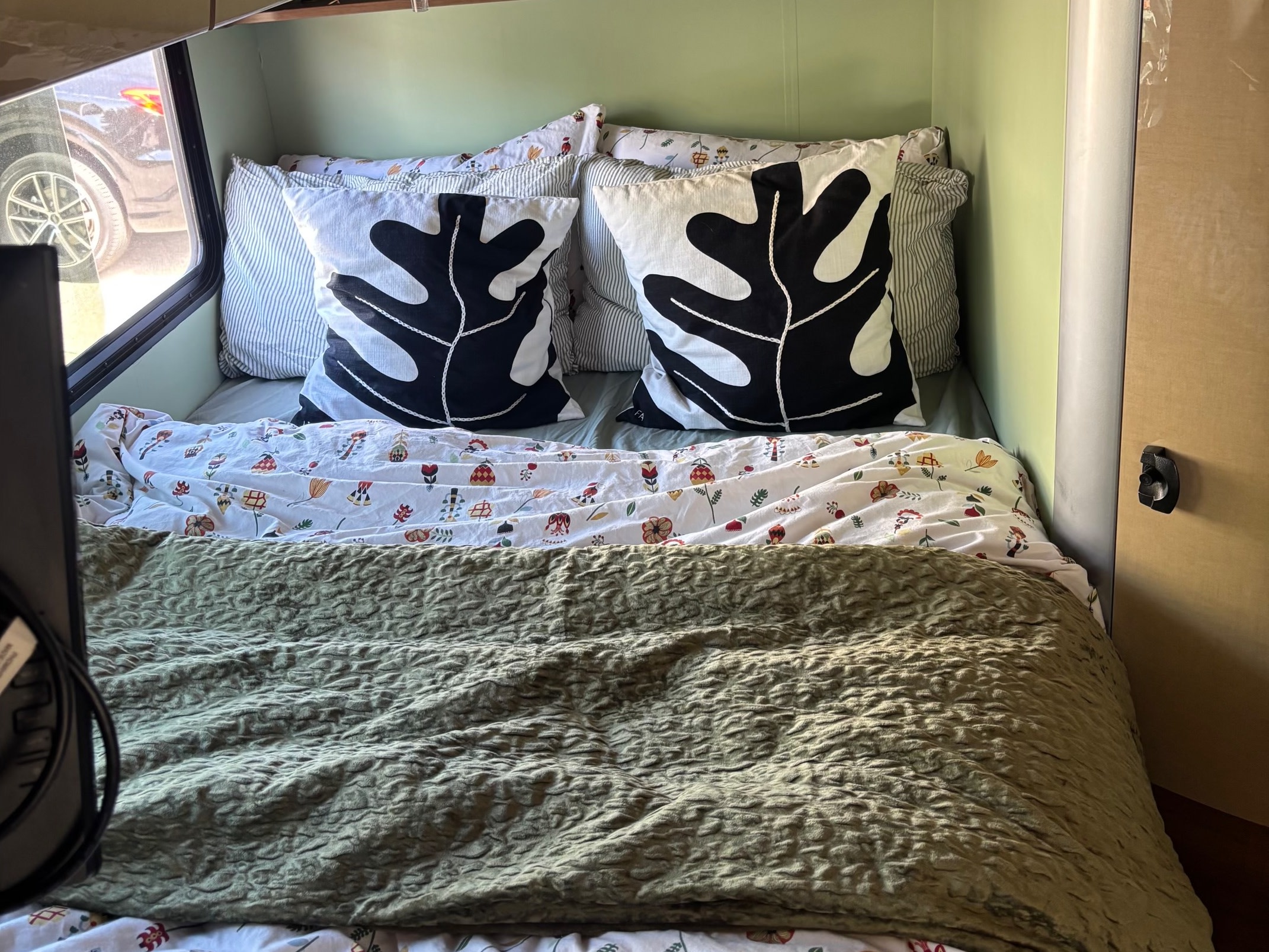 RV rental bedroom: a cozy bed with black leaf pillows, floral duvet, and a textured green blanket against light green walls.. Winnebago View 2016