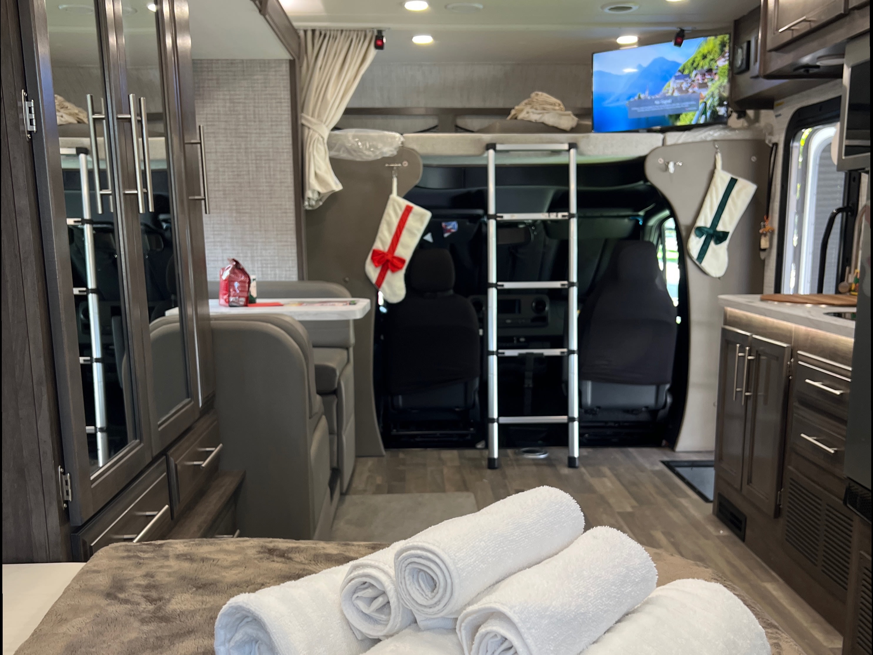 Inside a comfortable RV rental: foreground bed with towels, mid-ground kitchen and dinette, and bunk over cab.. Entegra Coach Qwest SE 2025