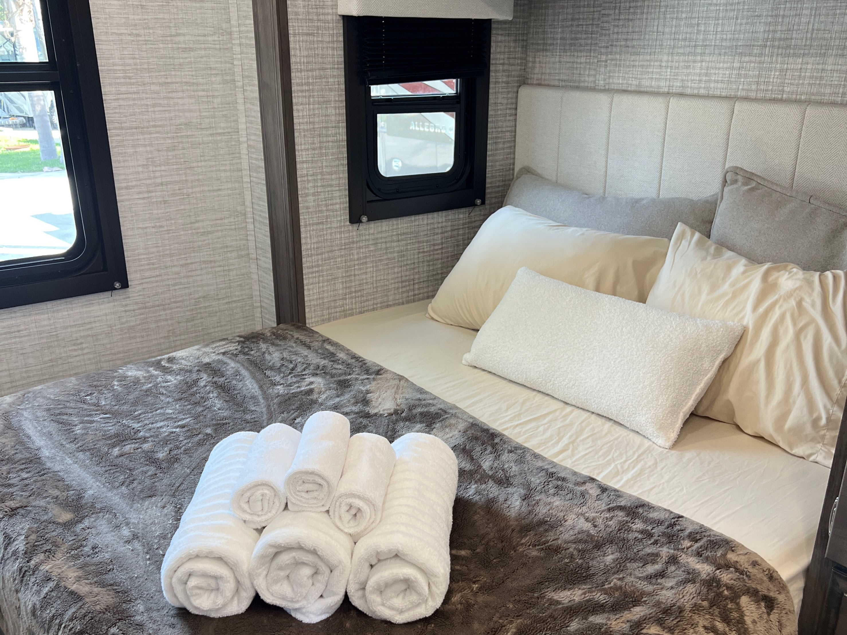 Inviting RV rental bedroom: plush bed with cream and grey pillows, soft blanket, and fresh rolled towels on a textured wall background.. Entegra Coach Qwest SE 2025