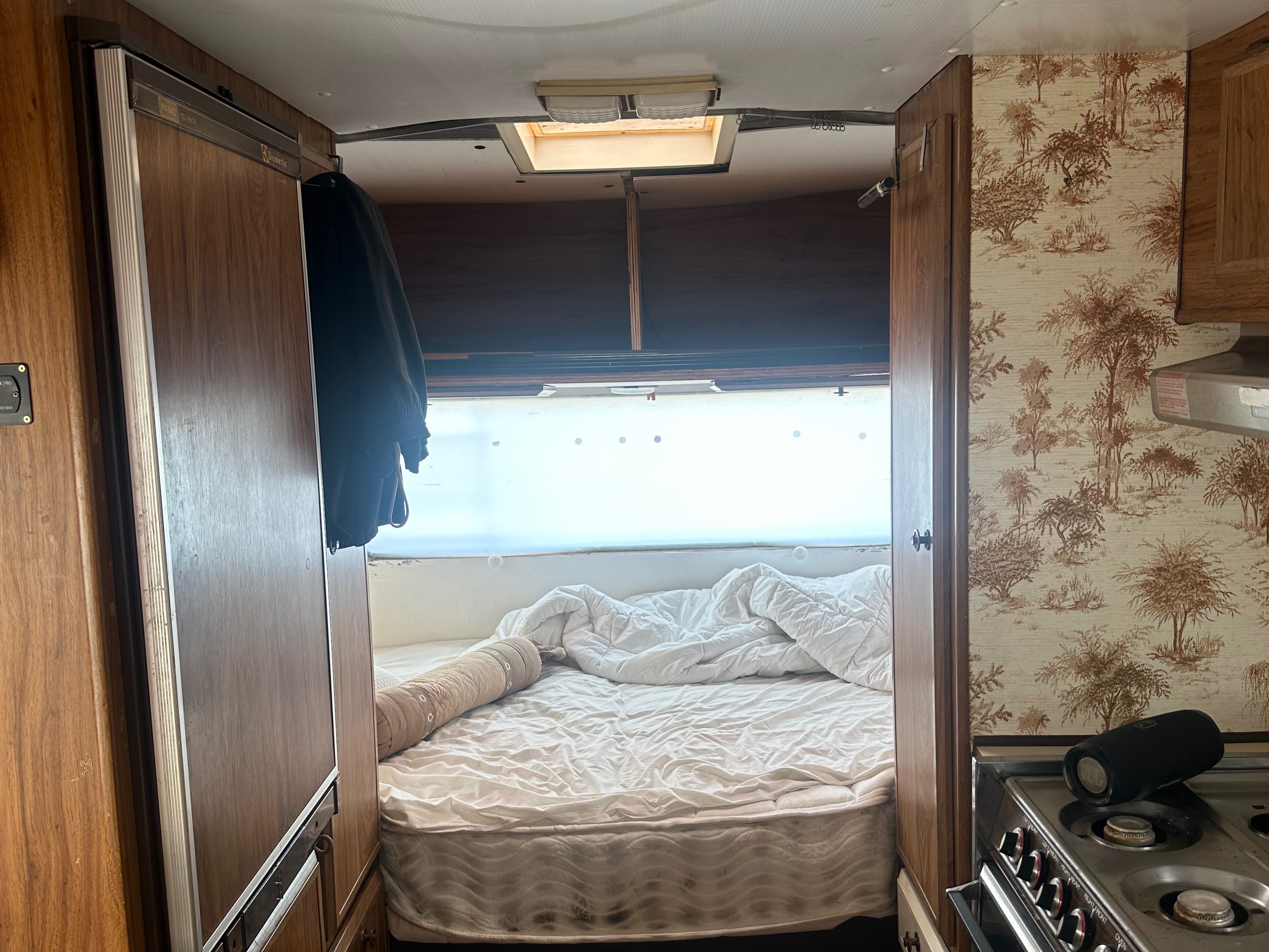Inside an rv rental, featuring a compact bedroom with a bed and a view into a vintage-style kitchen with a stove and fridge.. Winnebago Winnebago 1980