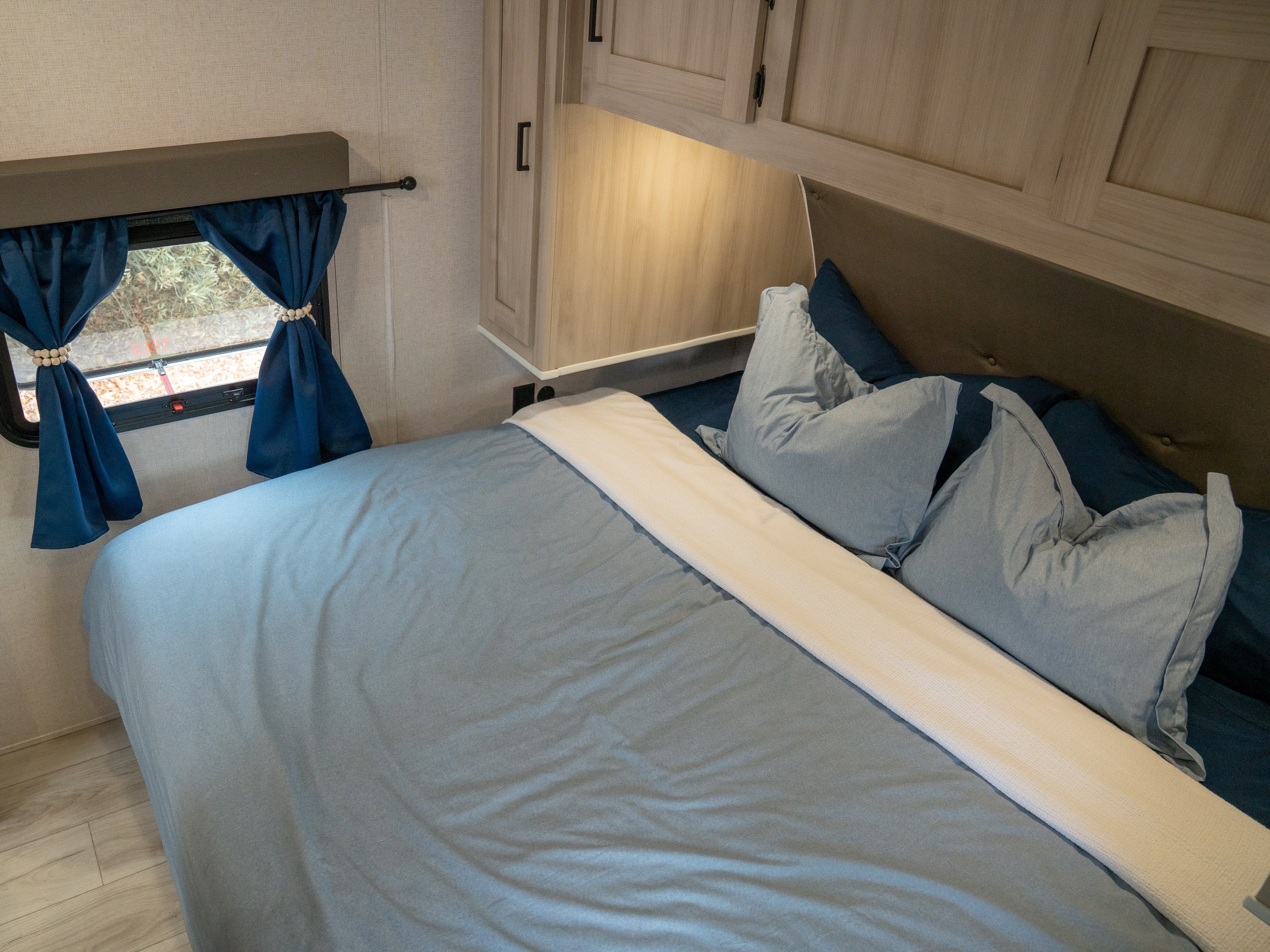 Cozy RV bedroom for an rv rental, featuring a light blue bed, wooden cabinets, and blue tie-back curtains next to a window.. East to West Della Terra 2025