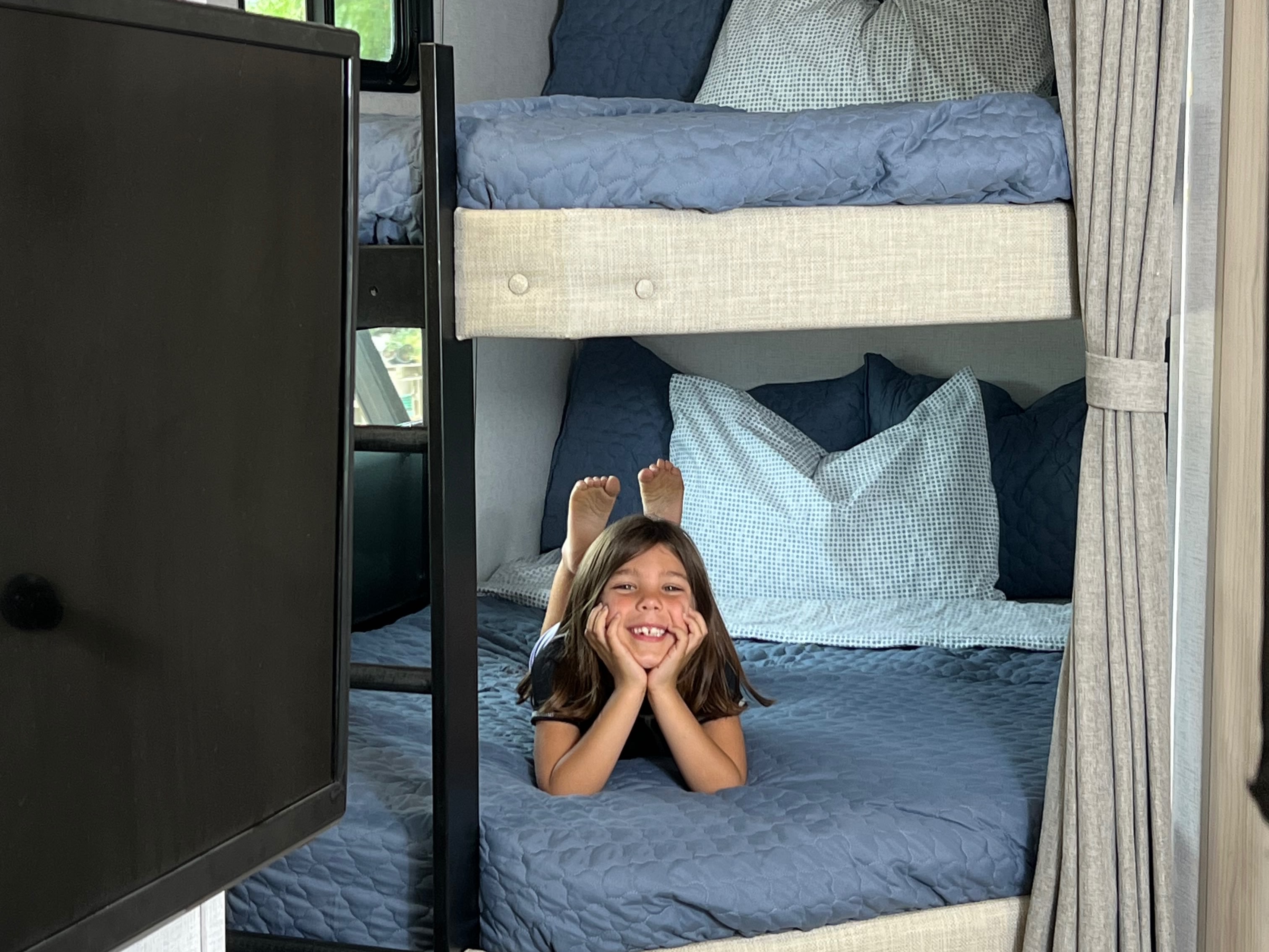 A happy girl smiles from a cozy blue bunk bed in an RV rental, ideal for family travel.. East to West Della Terra 2025