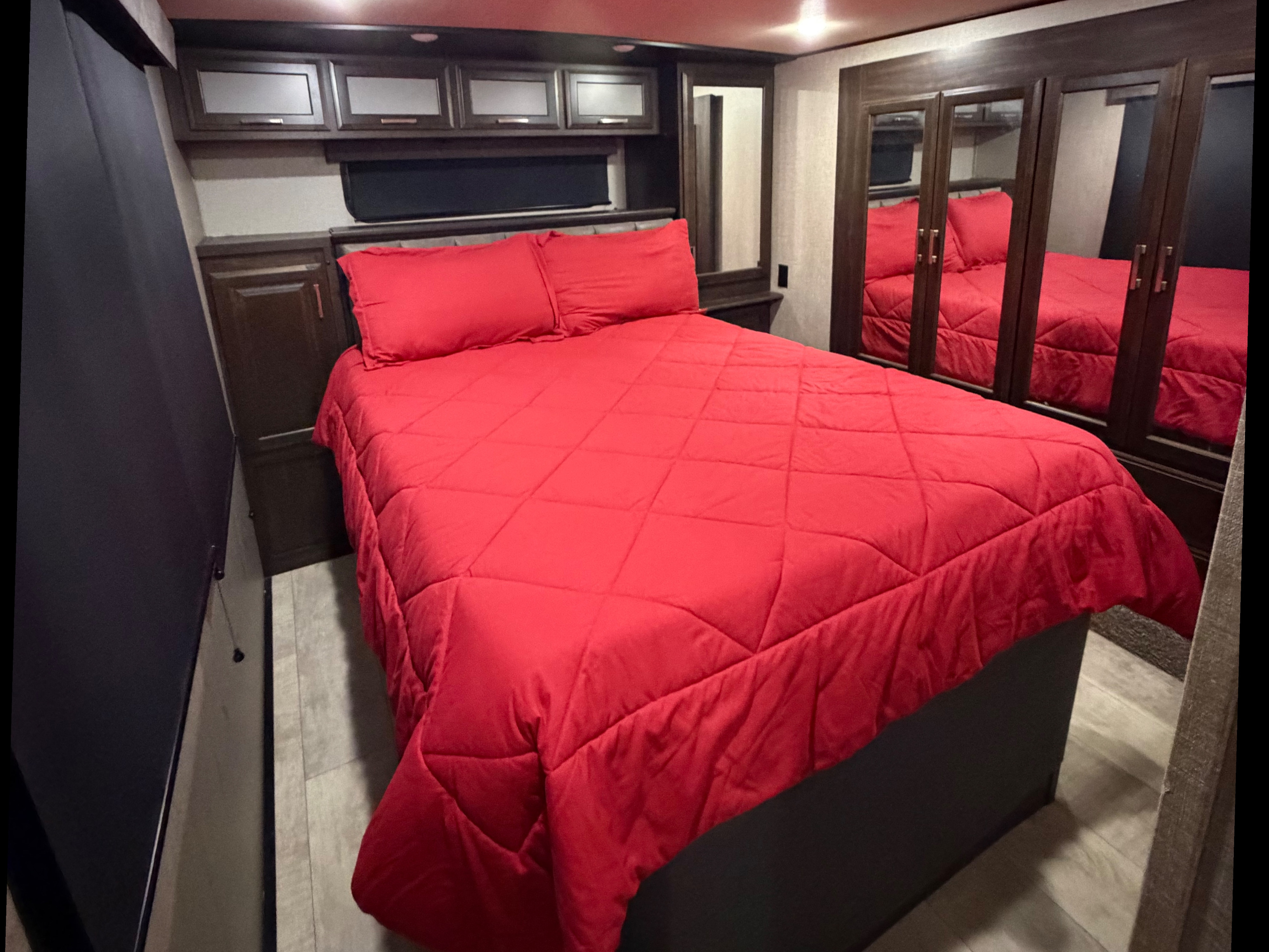 Inside an RV rental: a comfortable bedroom featuring a red quilted comforter, pillows, dark wood cabinets, and mirrored closet doors.. Grand Design Momentum Toy Hauler 2020