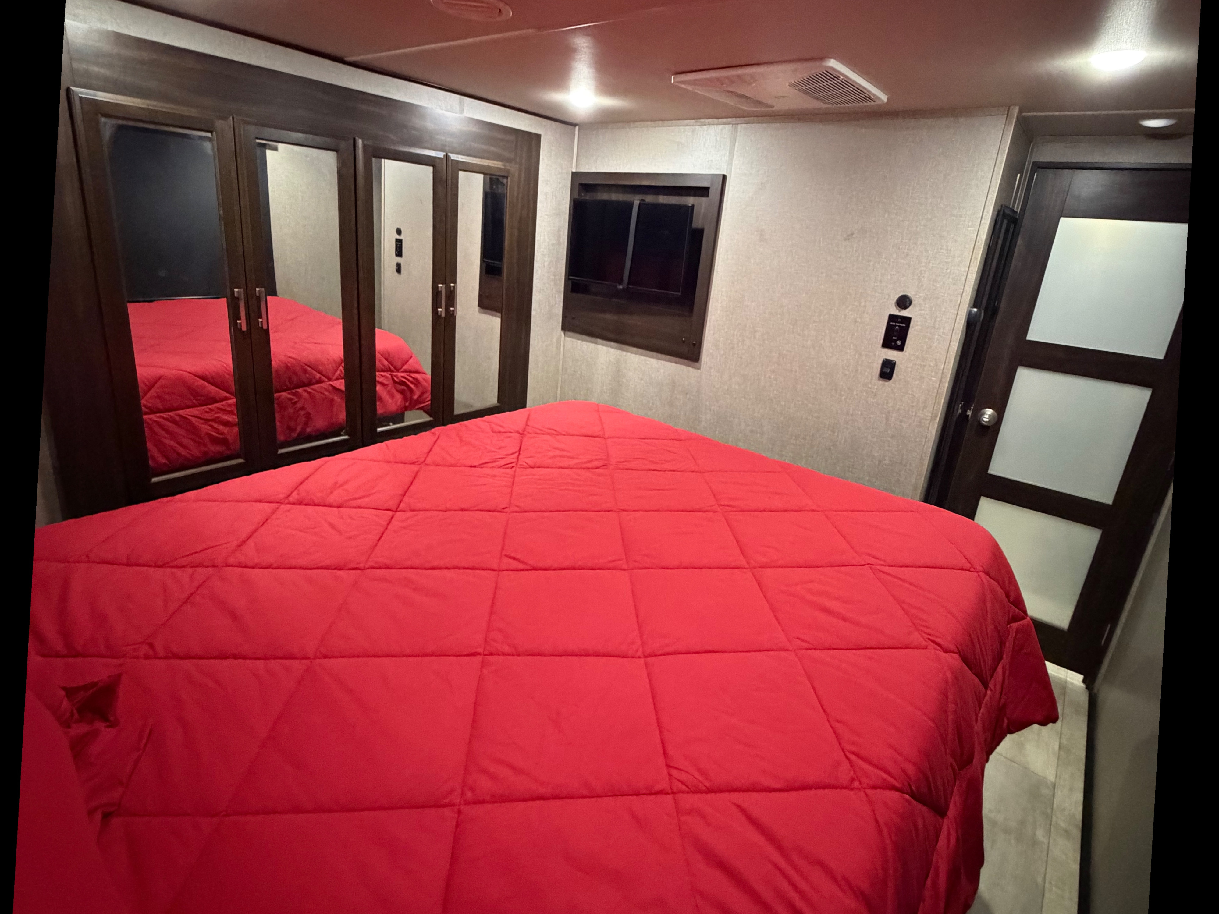 This RV rental bedroom features a comfy red bed, mirrored closet doors, a wall-mounted TV, and an overhead AC unit.. Grand Design Momentum Toy Hauler 2020