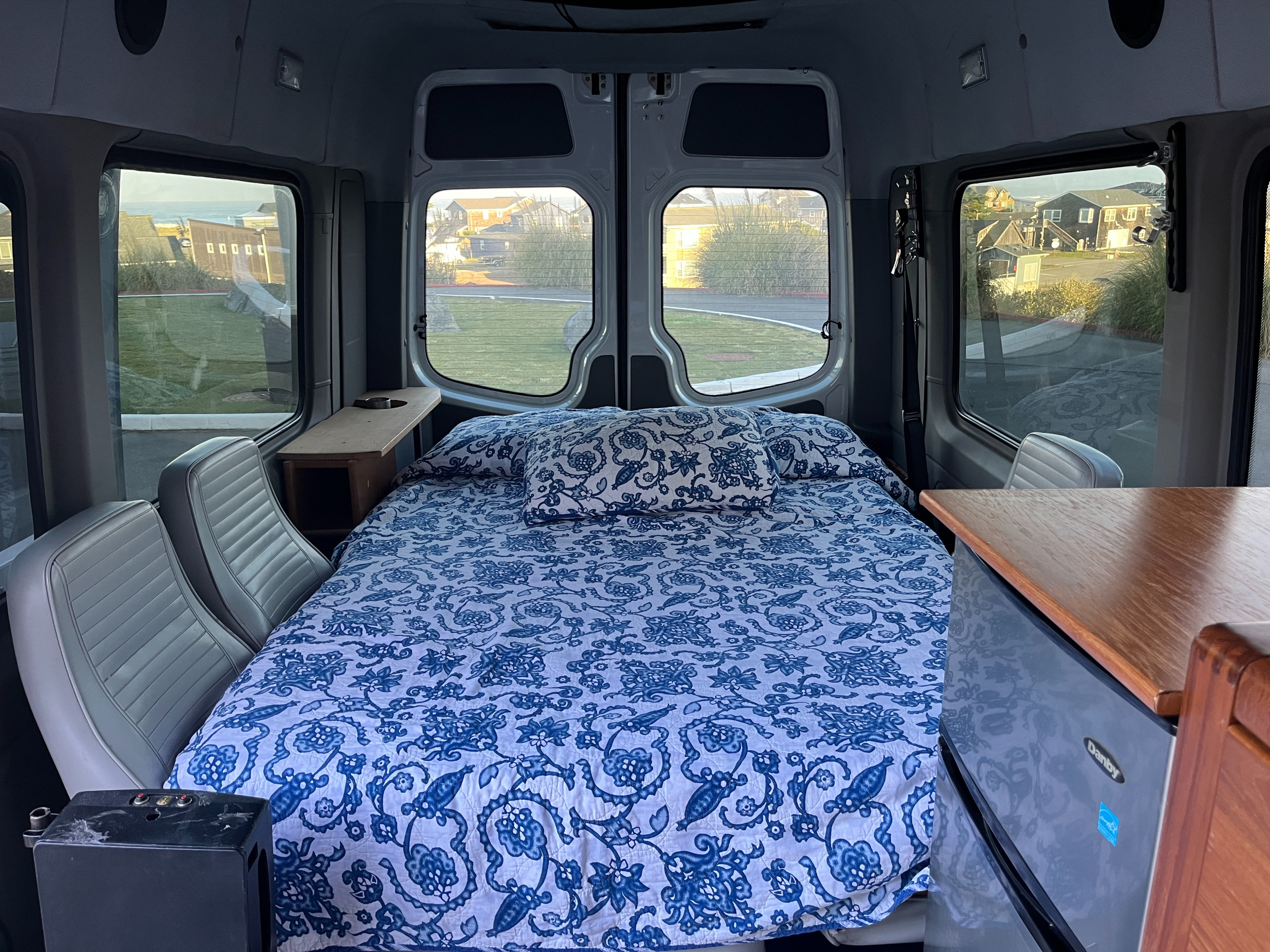 Inside a comfortable rv rental camper van: full bed with patterned bedding, mini-fridge, and scenic ocean views.. Winnebago Mercedes Sprinter 2014