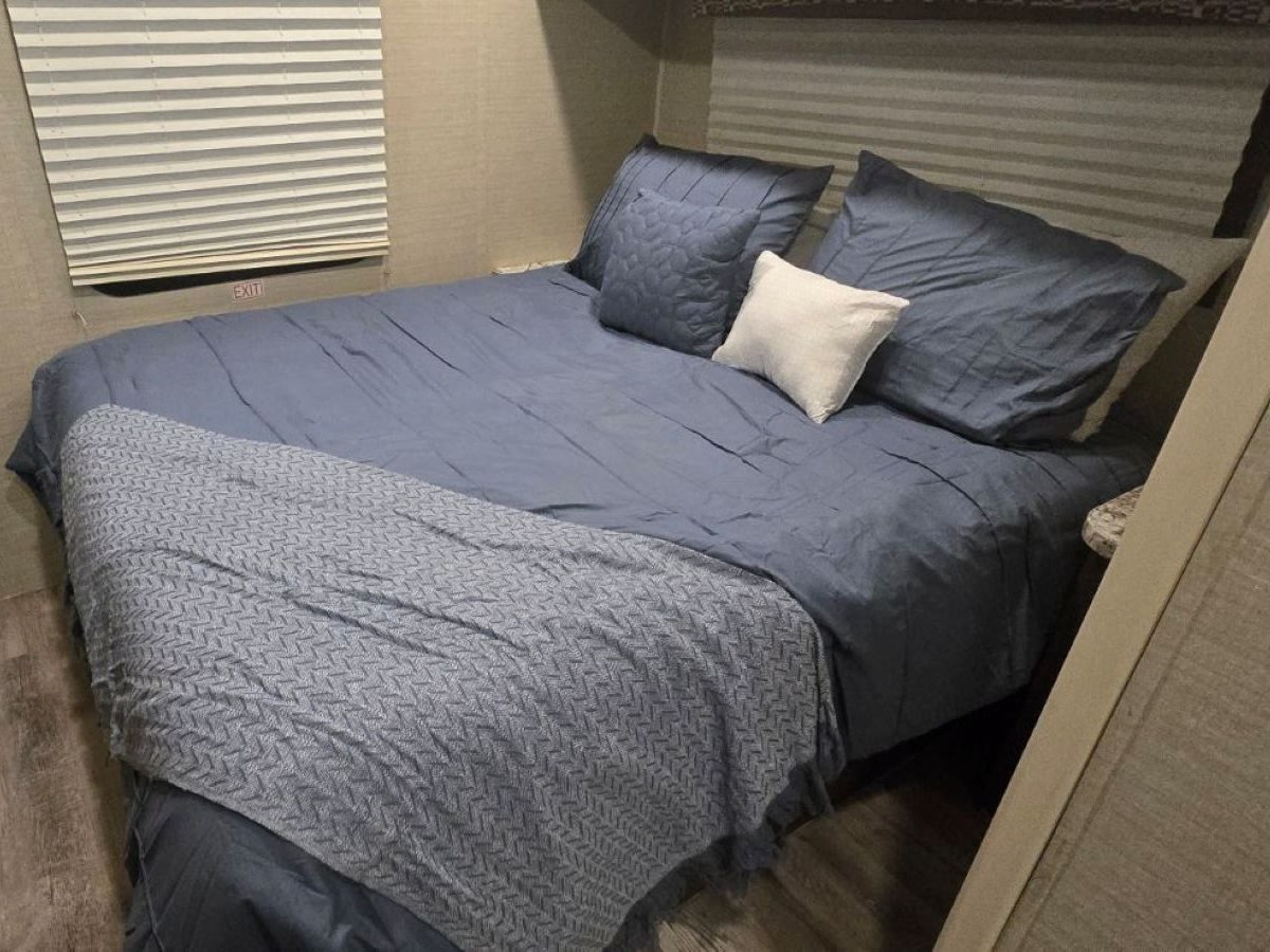 Comfortable RV rental bedroom with a blue bed, pillows, patterned throw blanket, and a window blind with an EXIT sign.. Jayco Redhawk 2019