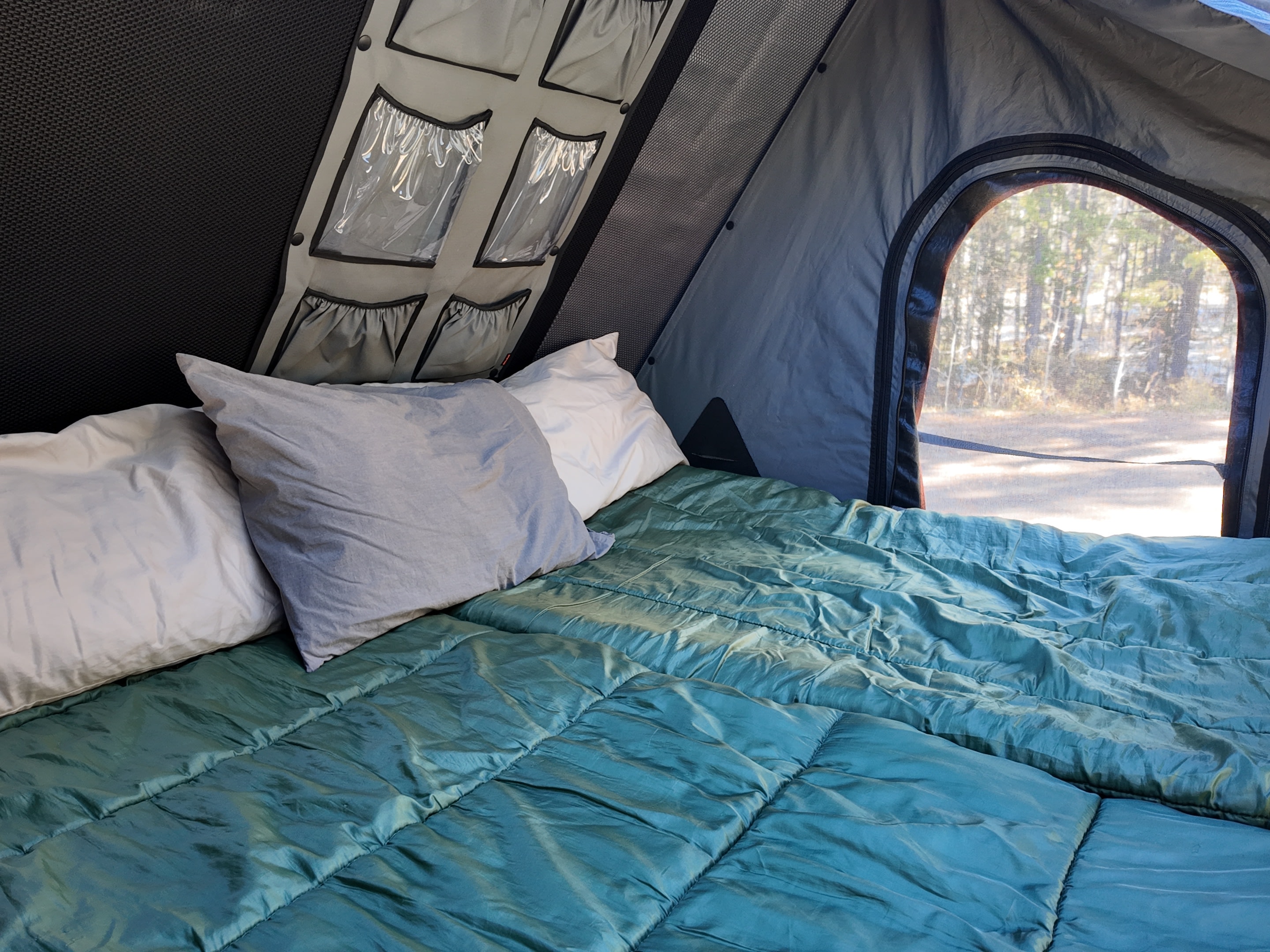 Cozy interior of an RV rental's roof top tent. A bed with two pillows and a teal comforter offers a forest view through the zipped window.. Ford Ranger 2300 Cc 2024
