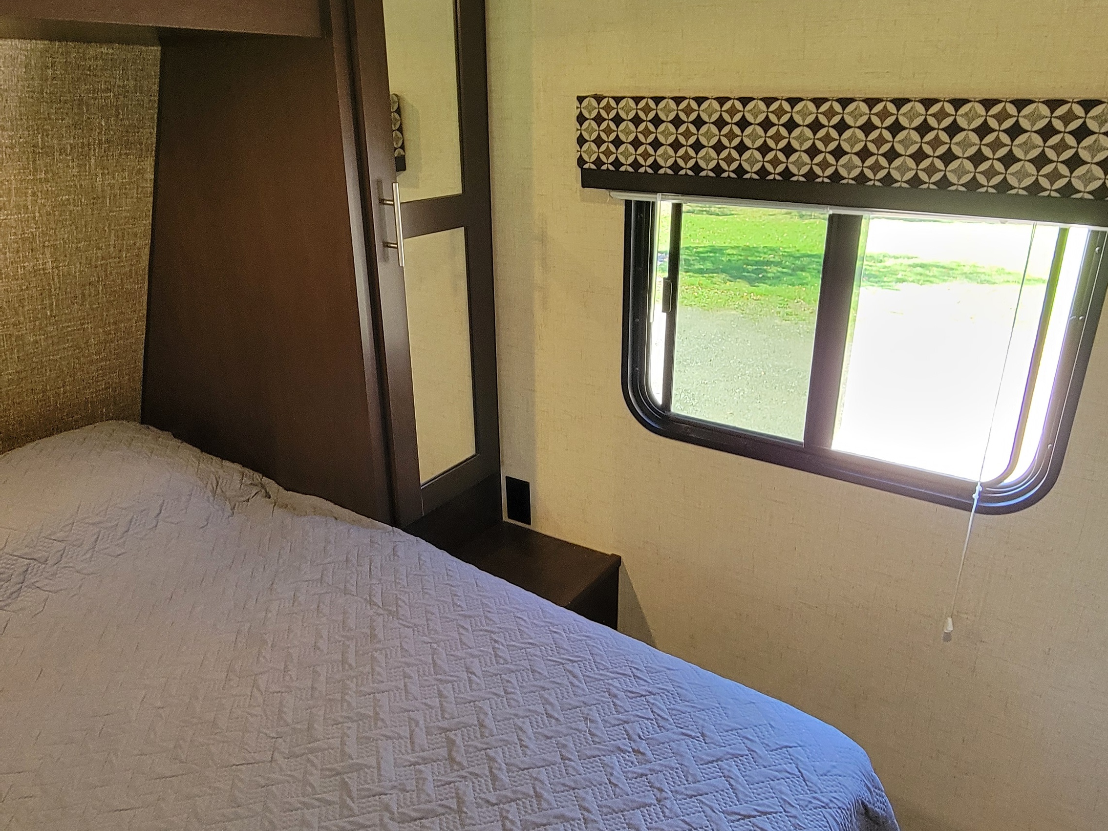 RV rental bedroom showing a gray quilted bed, dark cabinet with mirror, small nightstand, and window with a geometric valance.. KZ 231RKLE 2018