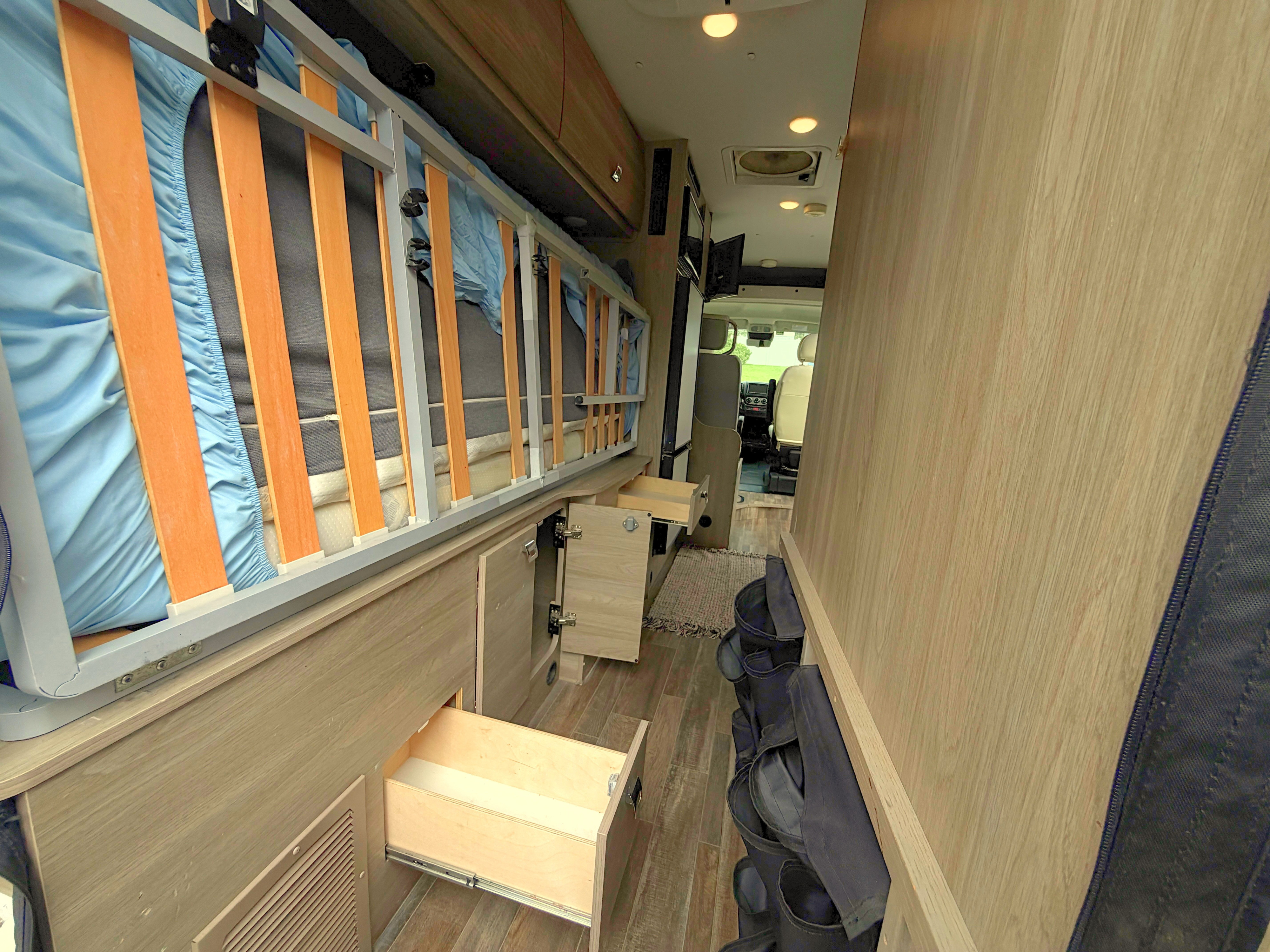 RV rental interior: elevated bed with blue sheet, open cabinets/drawers, and shoe storage in a narrow aisle towards the front cabin.. Winnebago Travato 2022