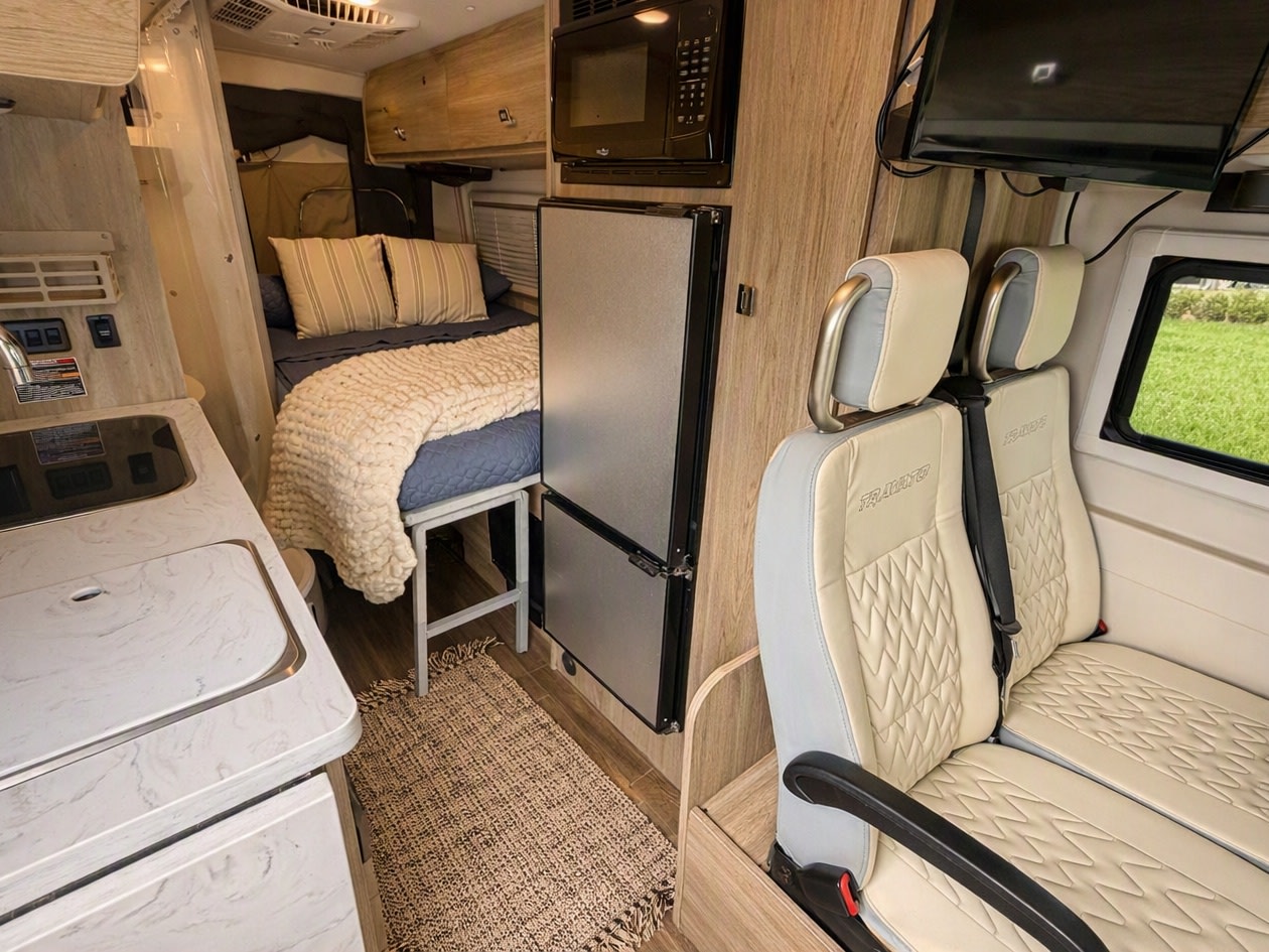 Cozy rv rental interior featuring a compact kitchen, bed with knit blanket, fridge, and passenger seats with TV.. Winnebago Travato 2022