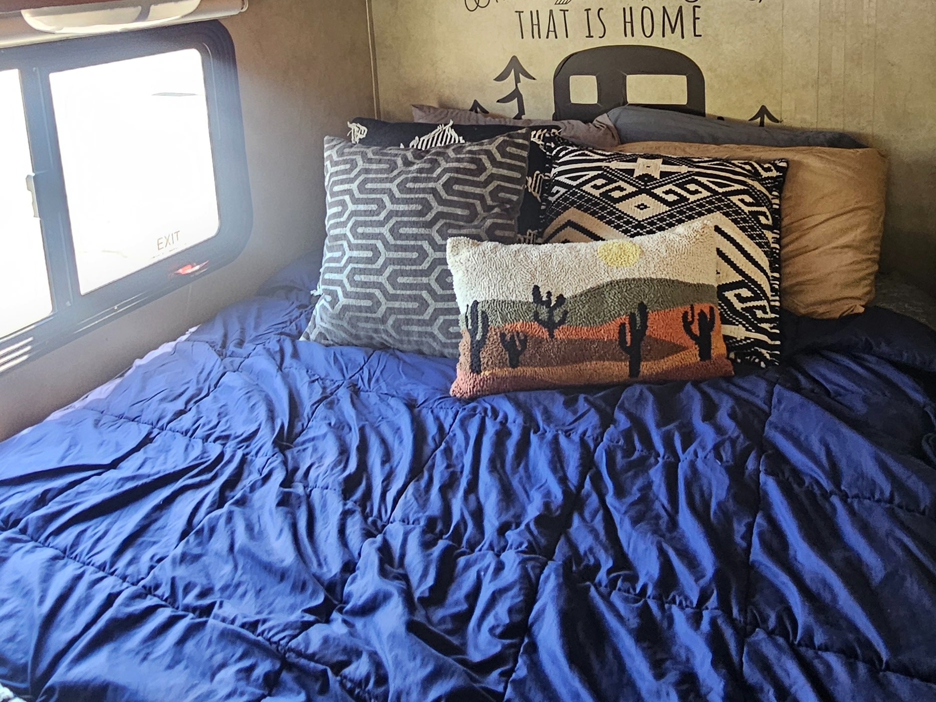 Cozy camper bedroom in an RV rental with blue bedding and desert-themed pillows. A "THAT IS HOME" decal decorates the wall.. Thor Majestic 2019